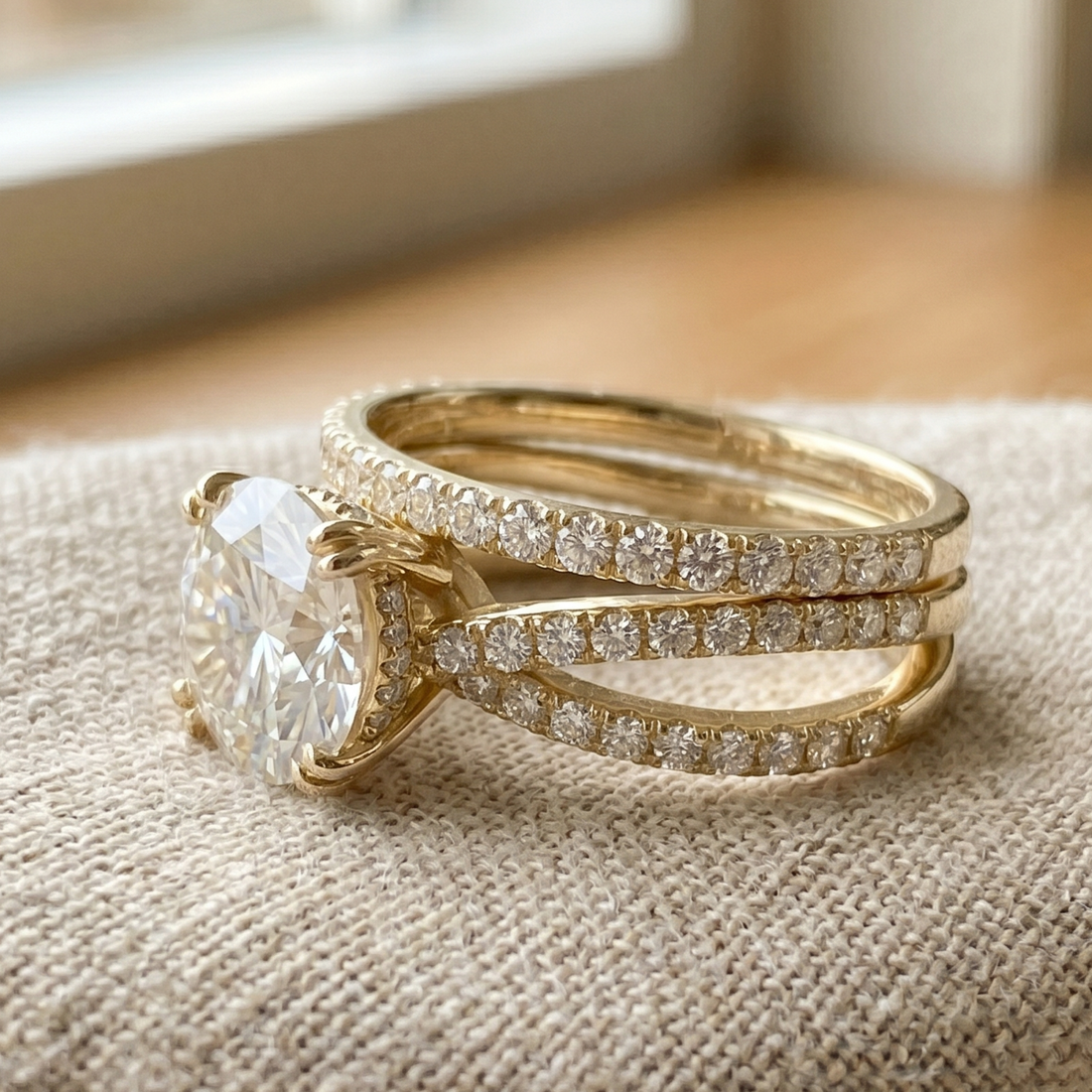 A gold wedding band set featuring a large oval-cut diamond solitaire and a matching pave diamond band, displayed on a textured beige fabric surface.