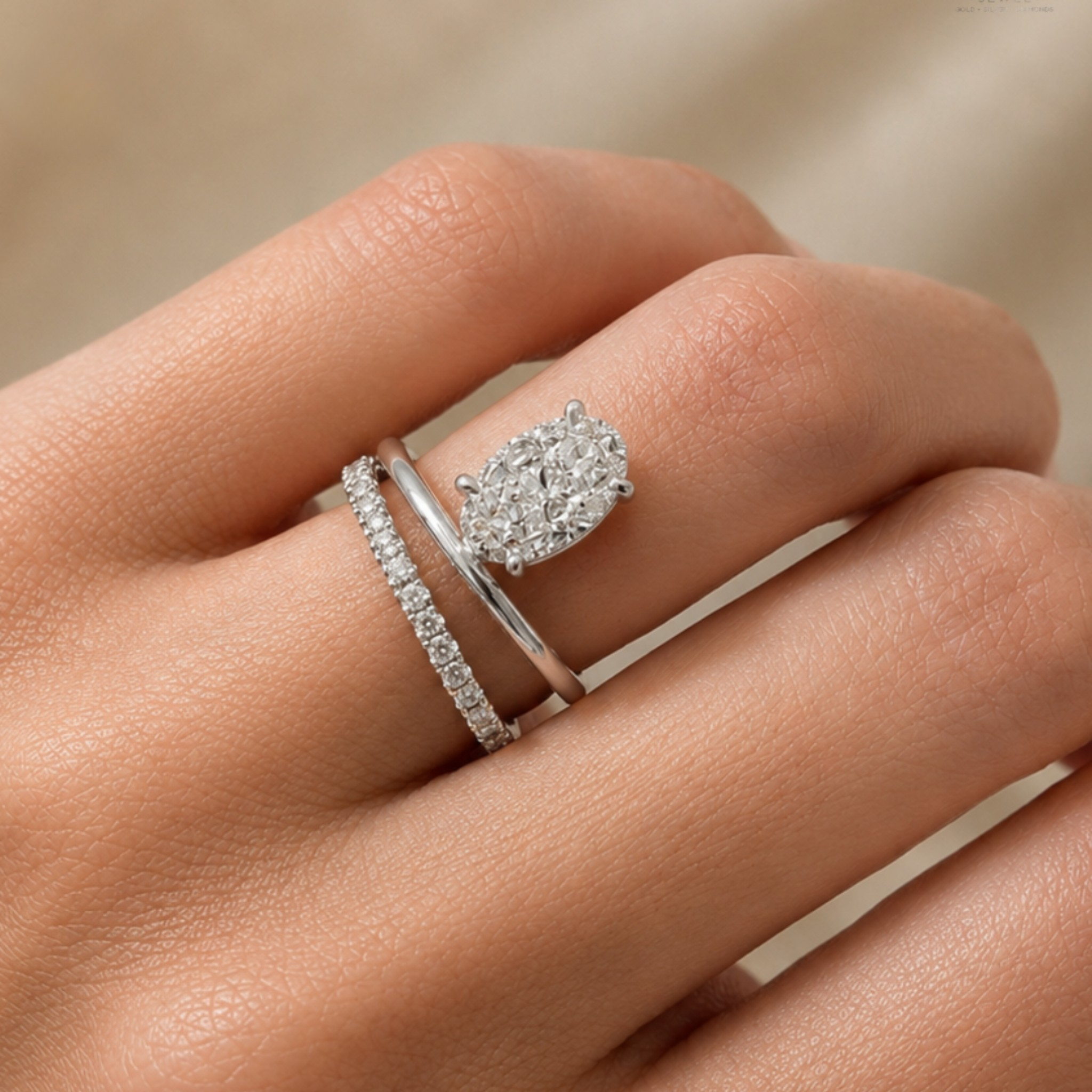 A close-up of a hand wearing two silver rings: one with a round cluster of clear stones and another with a row of small clear stones, against a soft beige background.