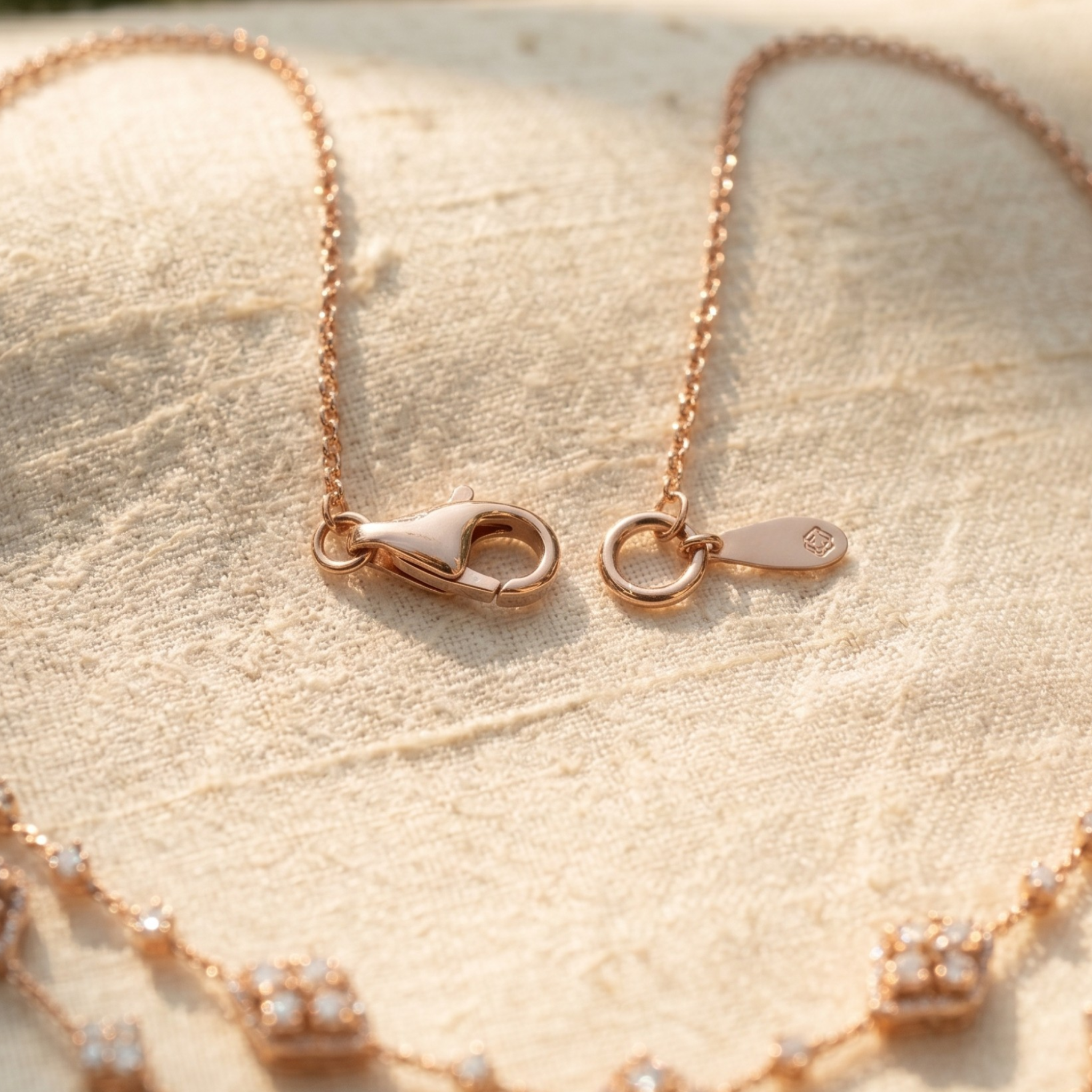 Rose gold chain necklace with a lobster clasp and a small oval tag, displayed on a textured beige fabric background.