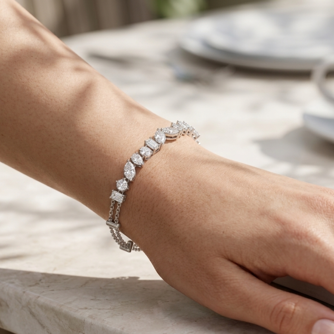 A close-up of a person's wrist wearing a silver-toned tennis bracelet with round and square-cut clear stones, set against a softly blurred outdoor table background.