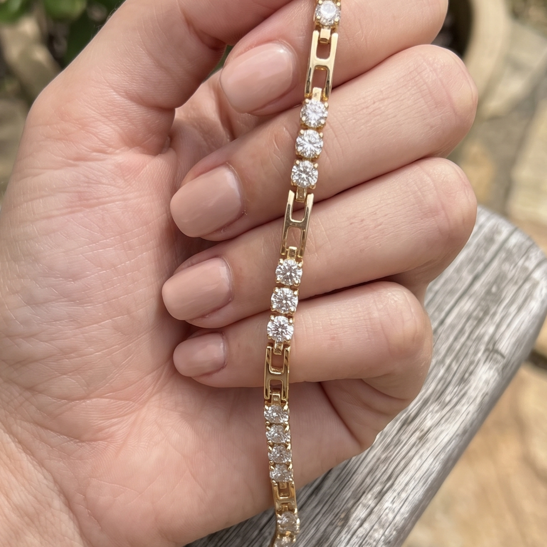 A gold tennis bracelet with round-cut clear stones, held in a person's hand against an outdoor wooden background.