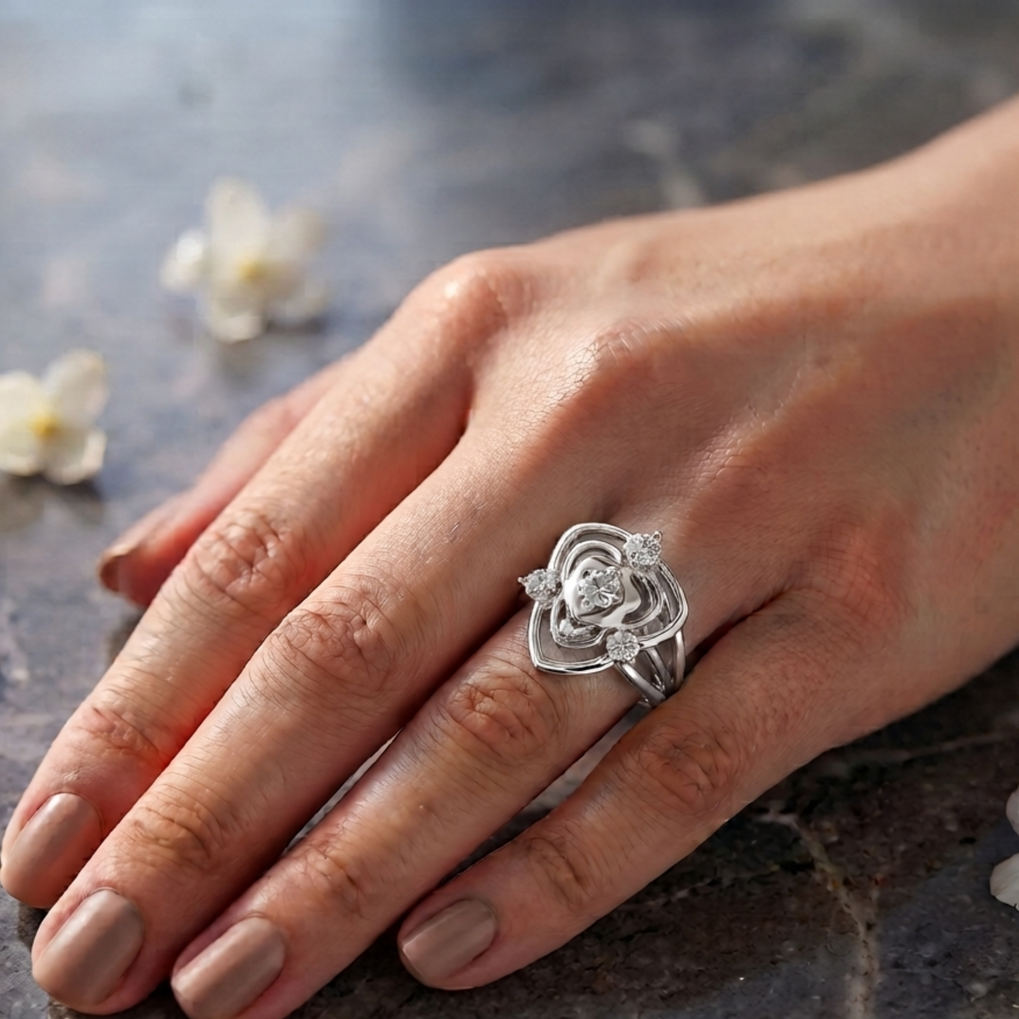 A silver ring with a sculpted rose design and small clear stones, worn on a woman's hand resting on a dark stone surface.