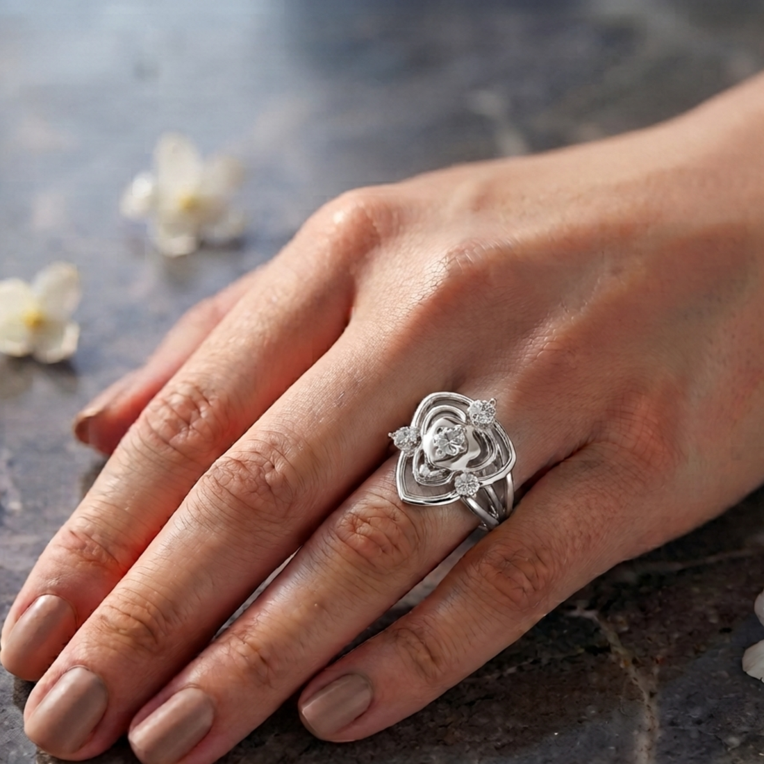 A silver ring with a sculpted rose design and small clear stones, worn on a woman's hand resting on a dark stone surface.