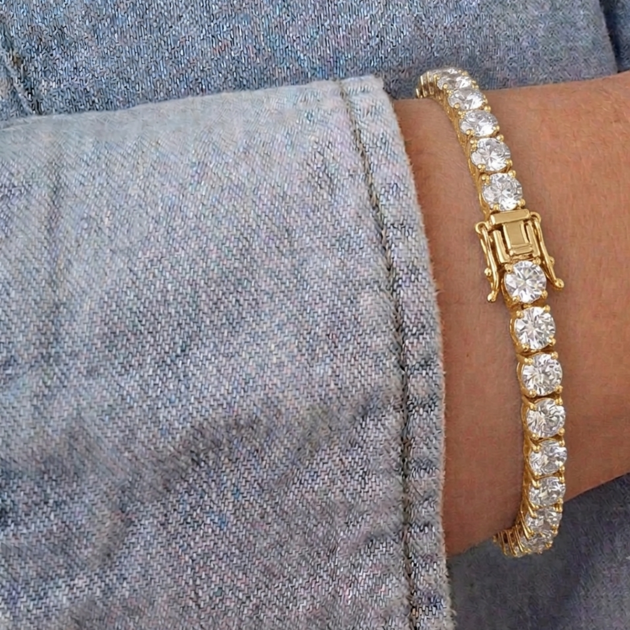 A close-up of a gold tennis bracelet with clear round-cut stones, worn on a wrist against a gray denim sleeve.