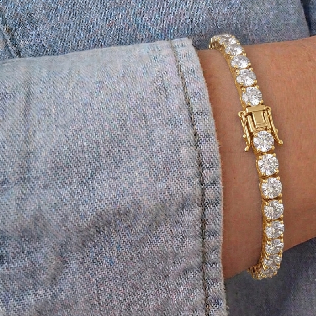 A close-up of a gold tennis bracelet with clear round-cut stones, worn on a wrist against a gray denim sleeve.