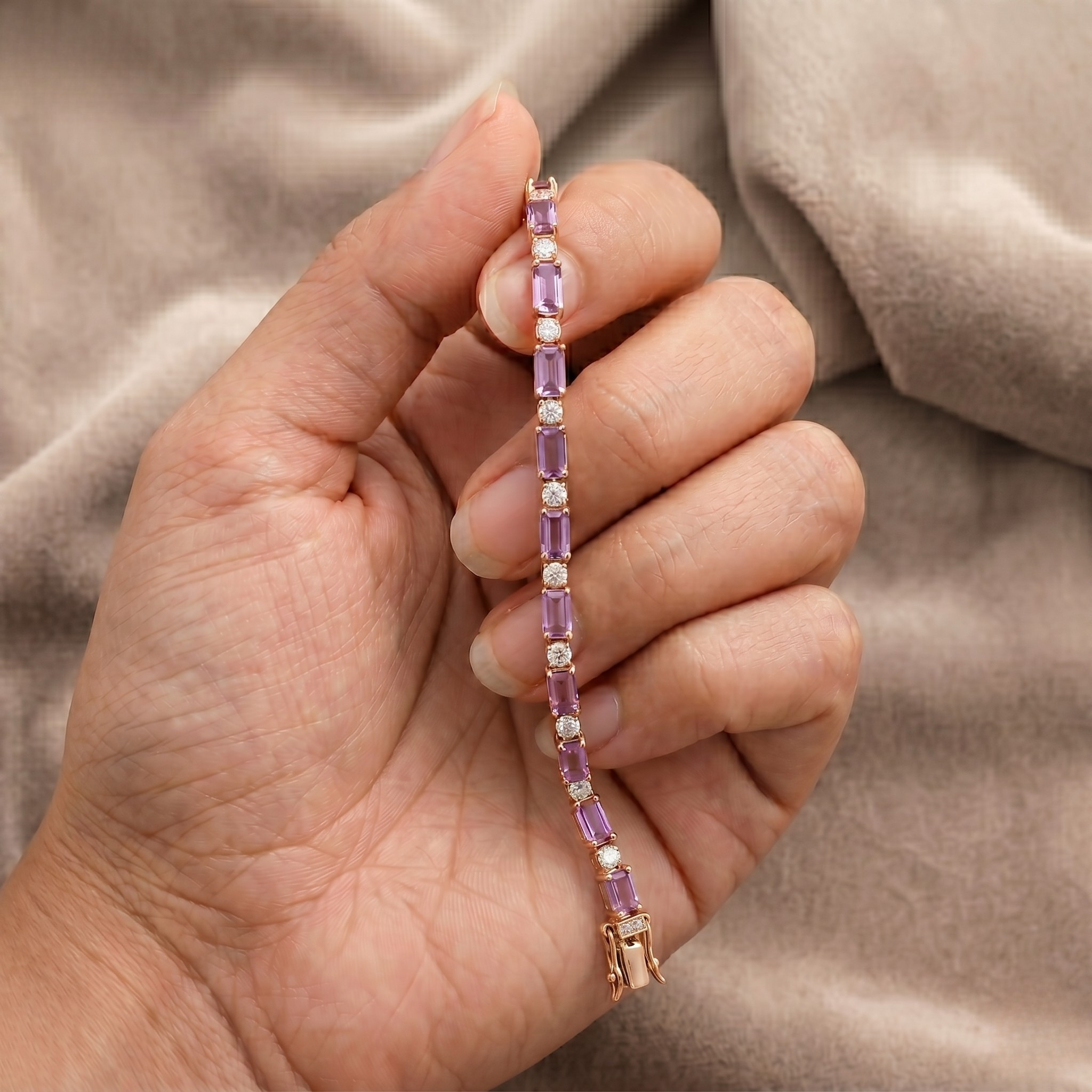 A close-up of a hand holding a delicate tennis bracelet with rectangular purple gemstones and small white stones set in rose gold, against a soft beige fabric background.