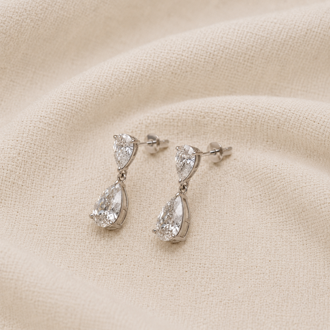 A pair of silver-tone drop earrings with pear-cut clear gemstones, displayed on a textured beige fabric background.