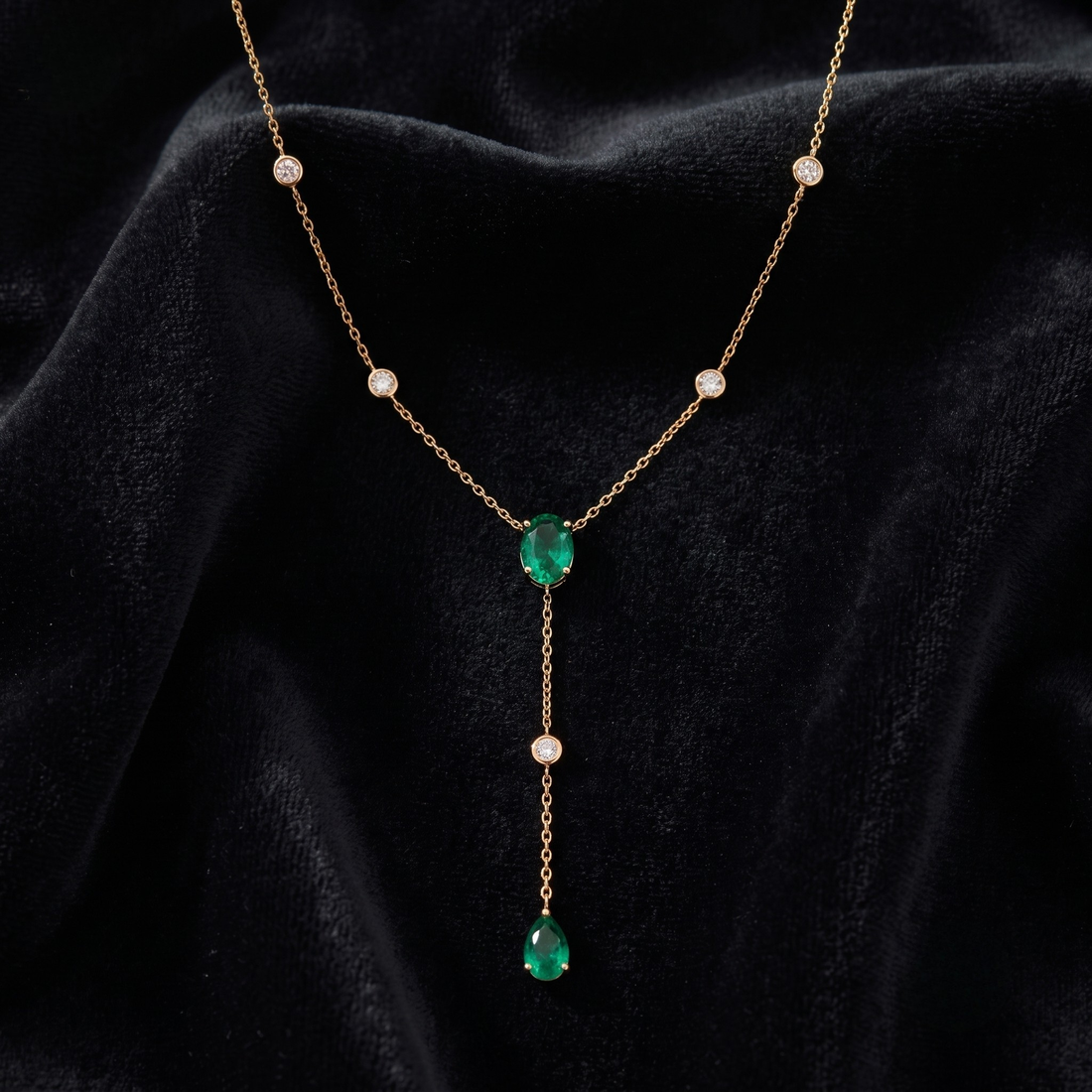 A delicate gold Y-necklace featuring two green emerald-cut center stones and five small clear accent stones, displayed on a draped black velvet fabric.