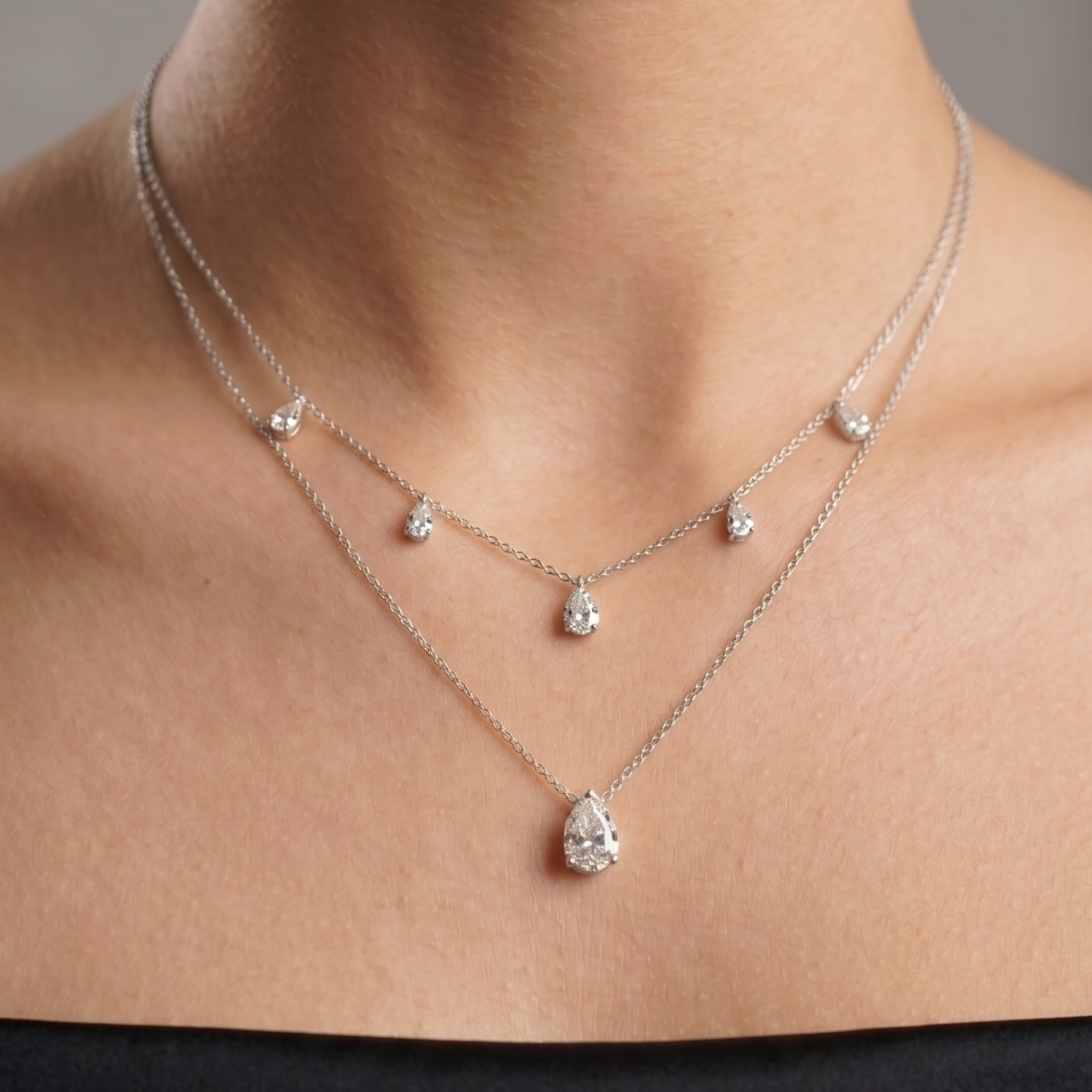 A close-up of a model's neck and upper chest wearing a delicate double-layered silver necklace with pear-cut clear gemstones.