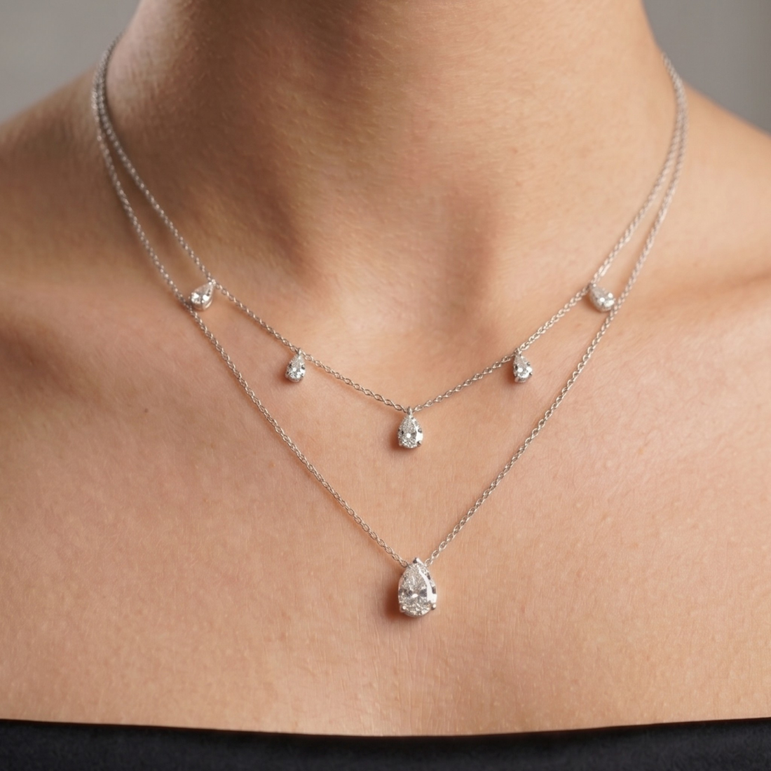 A close-up of a model's neck and upper chest wearing a delicate double-layered silver necklace with pear-cut clear gemstones.