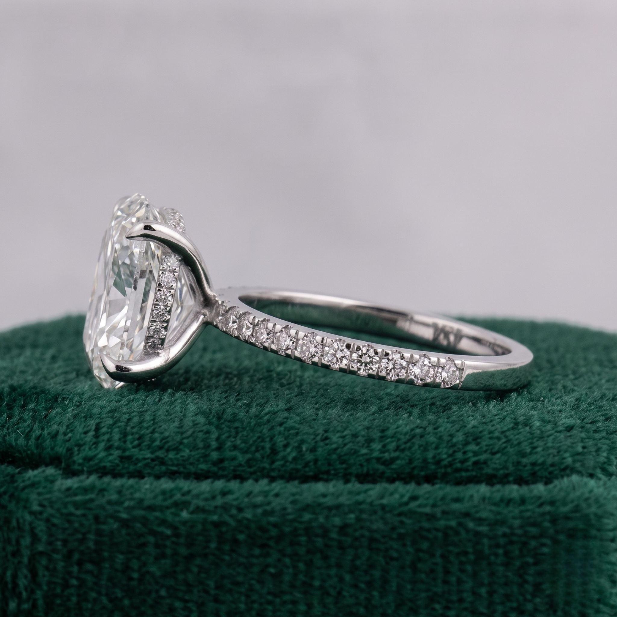 A silver-toned diamond engagement ring with a large oval-cut center stone and a pavé-set band, displayed on a textured dark green velvet ring box against a soft gray background.
