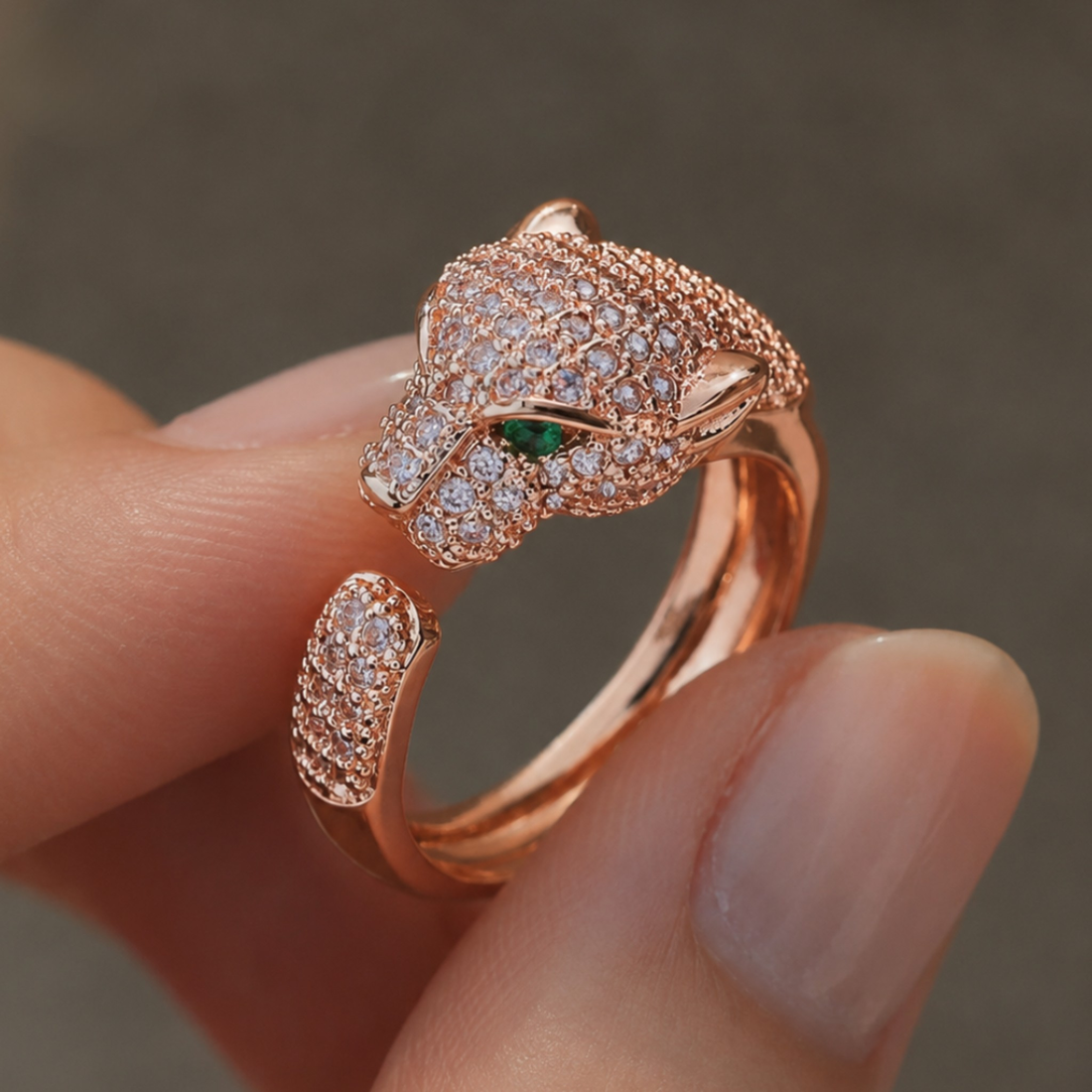A close-up of a rose gold open-band ring shaped like a panther head, set with pavé diamonds and a small green emerald accent in the eye, being worn on a person's fingers.