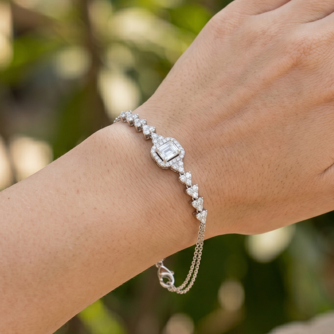 A silver-toned diamond bracelet with a central square-cut diamond and heart-shaped pavé accents, shown worn on a person's wrist against a blurred green background.