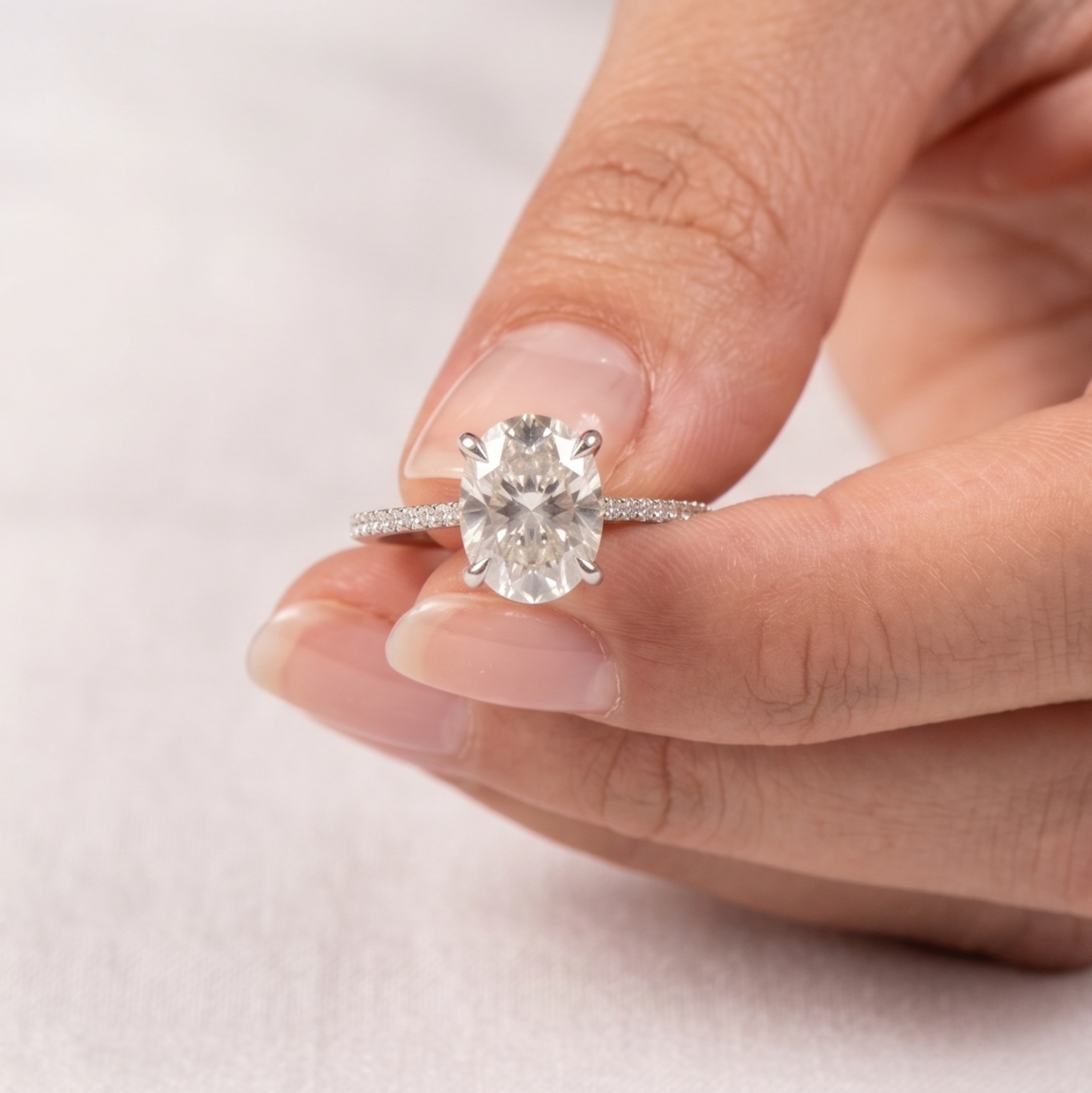 A close-up of a hand holding a silver engagement ring with a large oval-cut center stone and a pavé-set band, against a soft white background.
