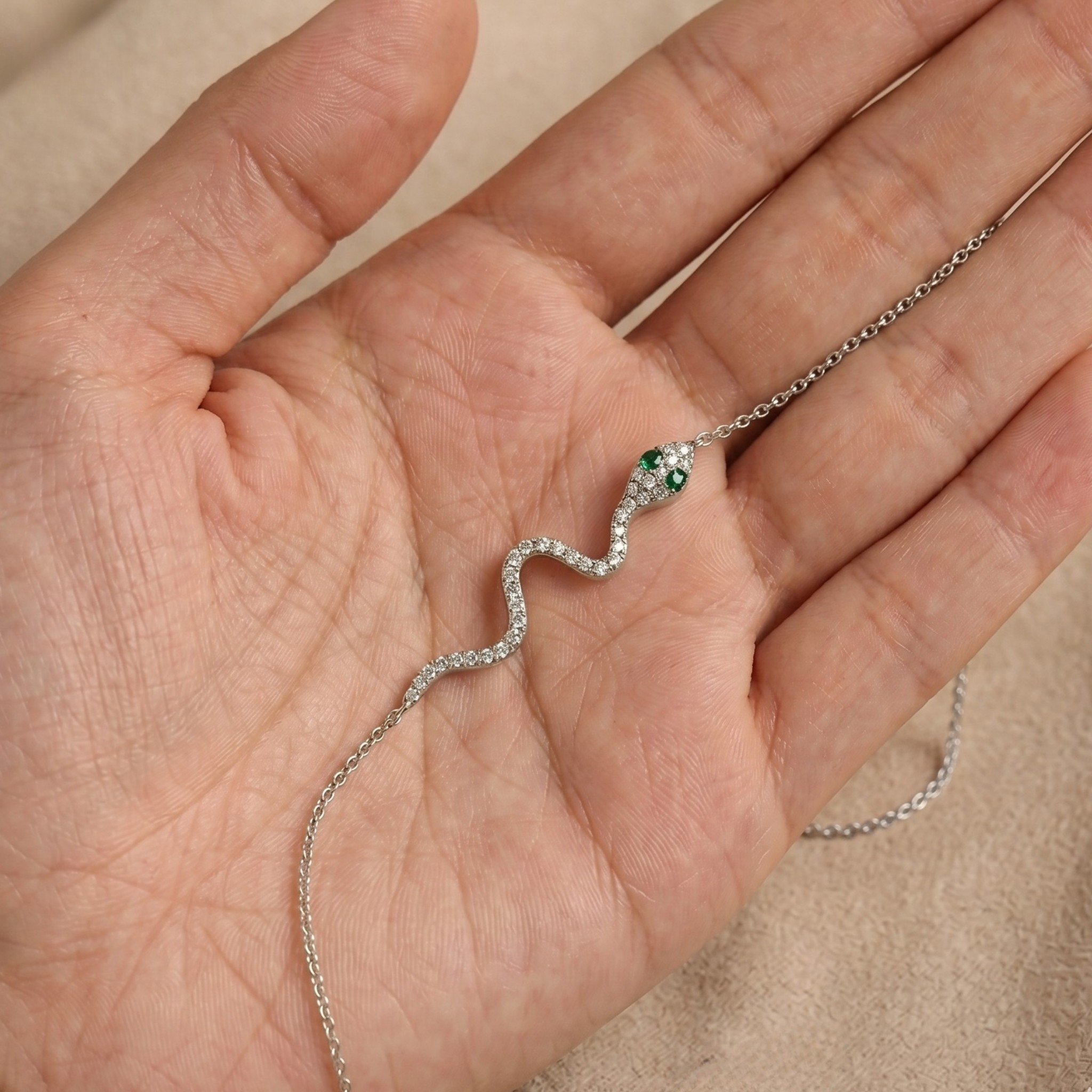A silver snake pendant necklace with green gemstone eyes and a diamond-encrusted body, displayed on a person's palm.