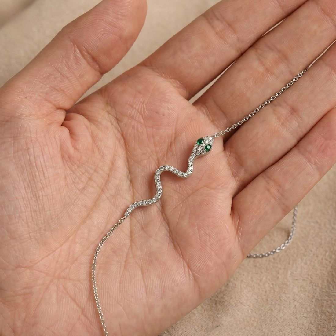 A silver snake pendant necklace with green gemstone eyes and a diamond-encrusted body, displayed on a person's palm.