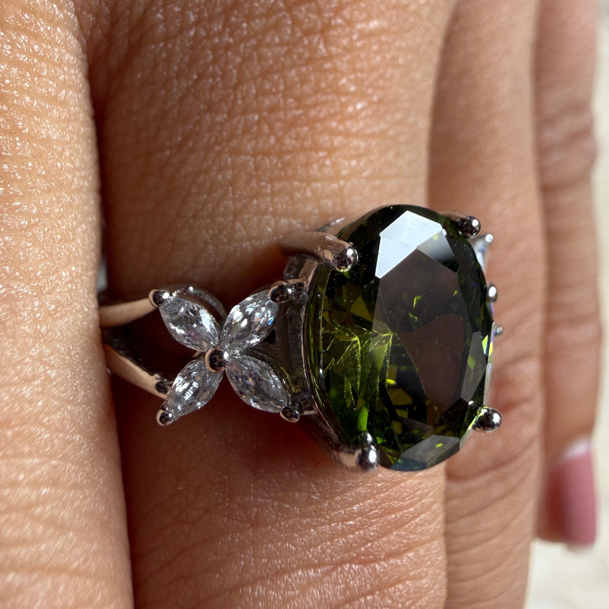 A close-up of a ring worn on a finger, featuring a large oval dark green gemstone in a silver-toned setting with marquise-shaped clear stones on the band.