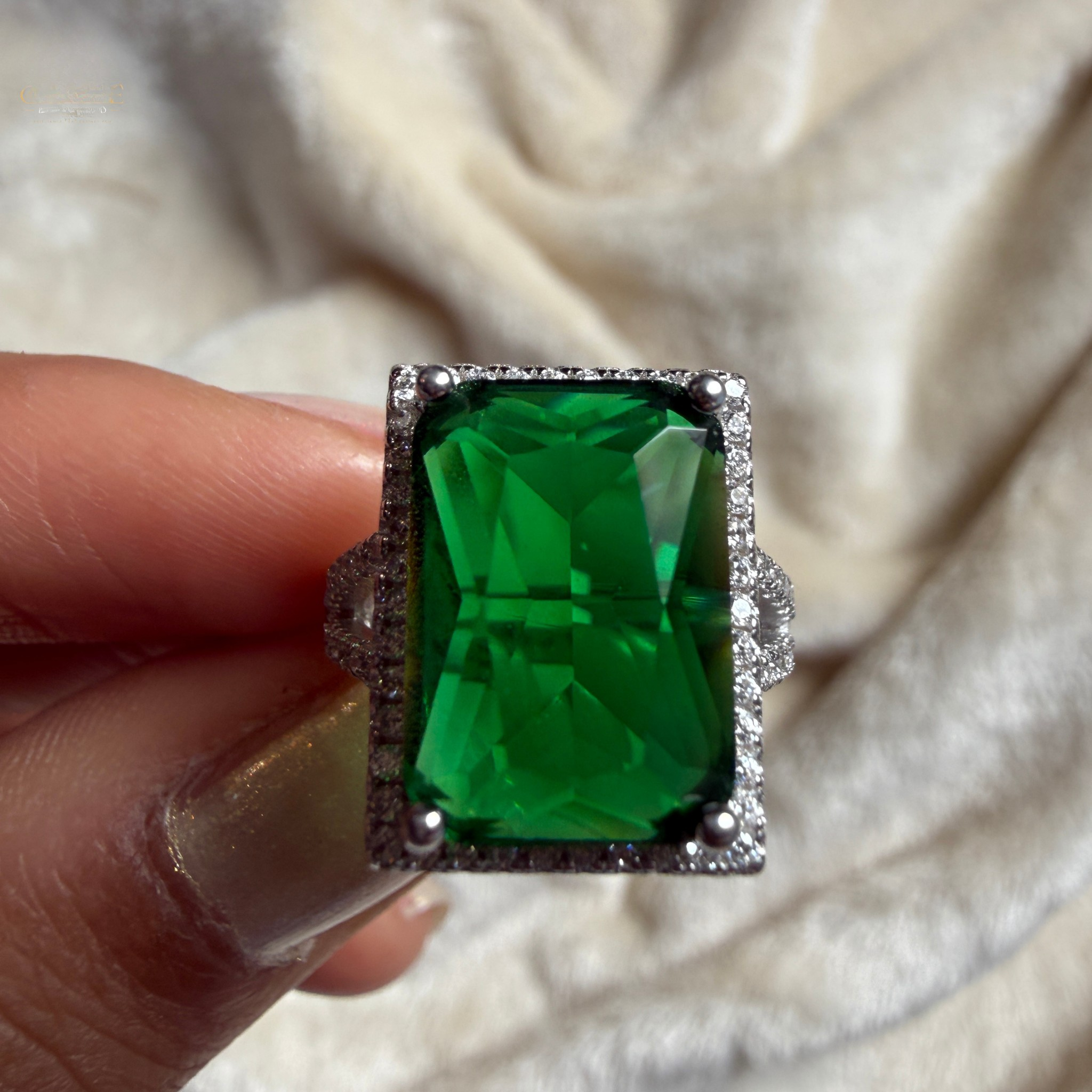 A rectangular emerald-cut green gemstone ring with a silver-toned halo setting, held between fingers against a soft cream-colored fabric background.