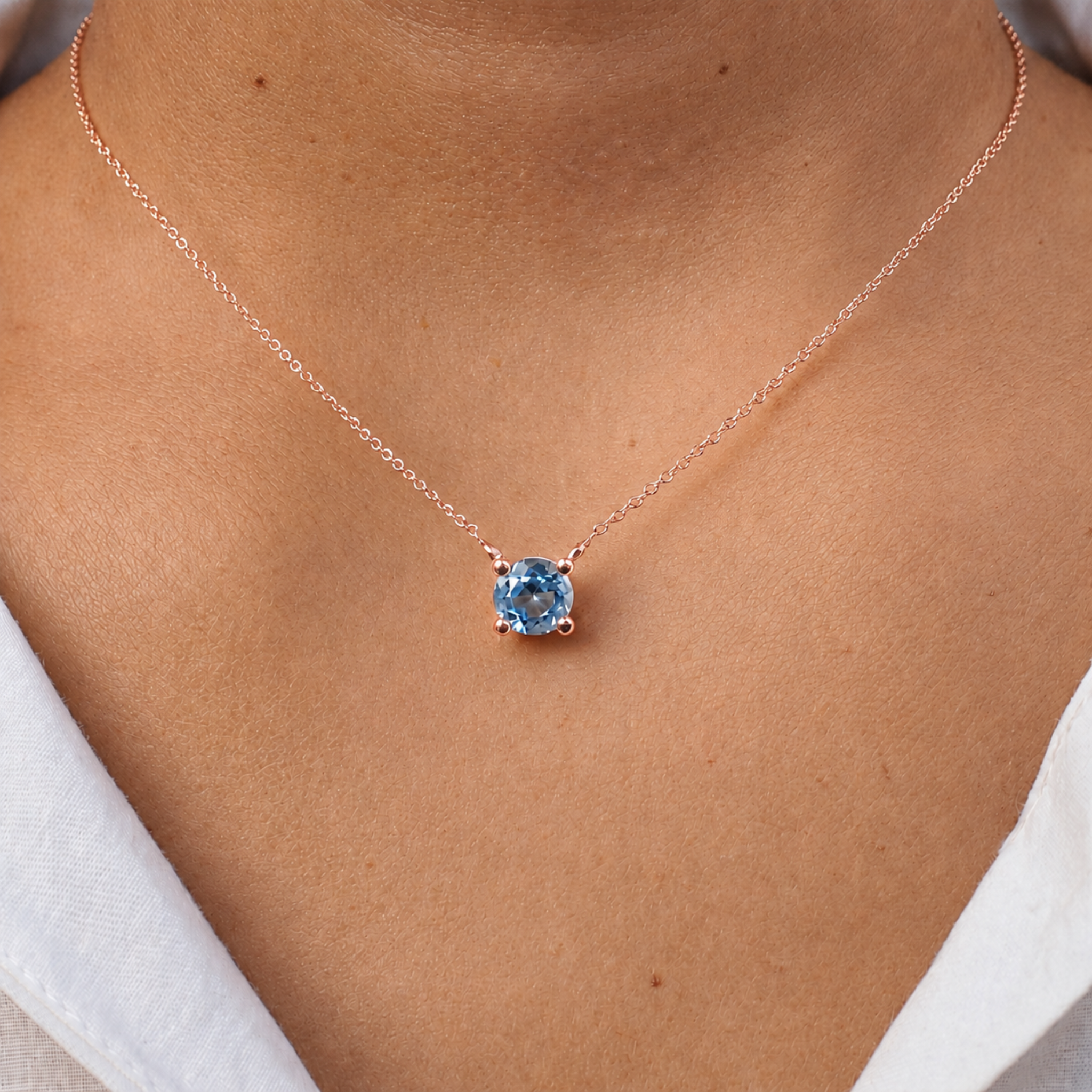 A close-up of a person's neck and chest wearing a delicate rose gold chain necklace with a round-cut light blue gemstone pendant.