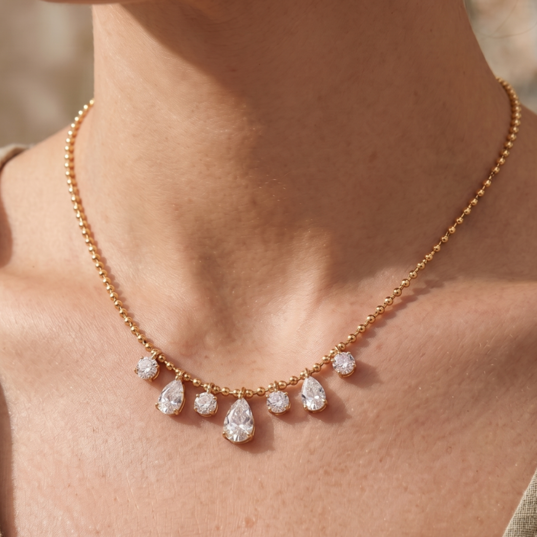 A close-up of a model's neck and chest wearing a delicate gold ball-chain necklace with a row of clear teardrop and round gemstones.