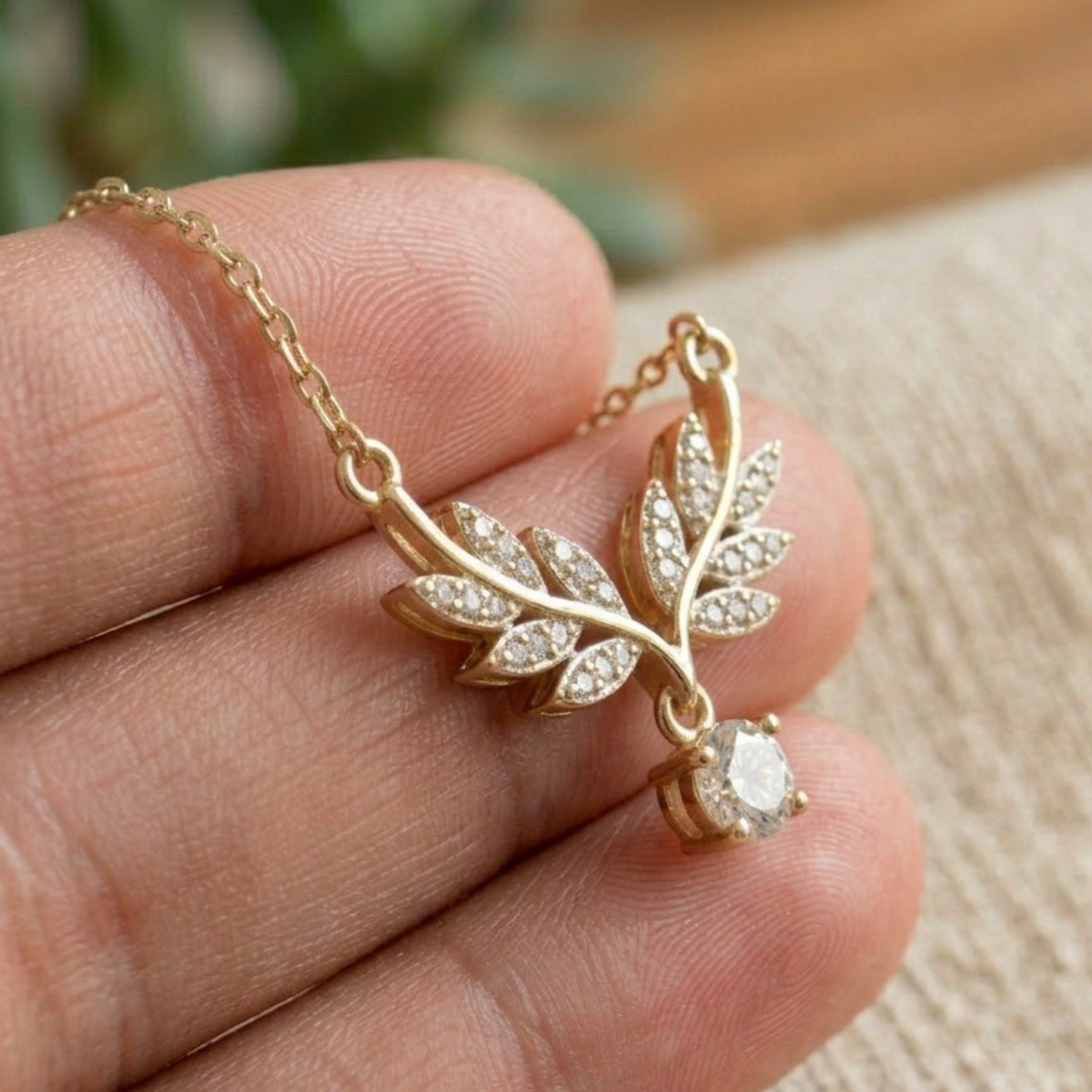 A gold leaf pendant necklace with clear pavé stones and a dangling round clear gemstone, held on a person's fingers against a soft beige fabric background.