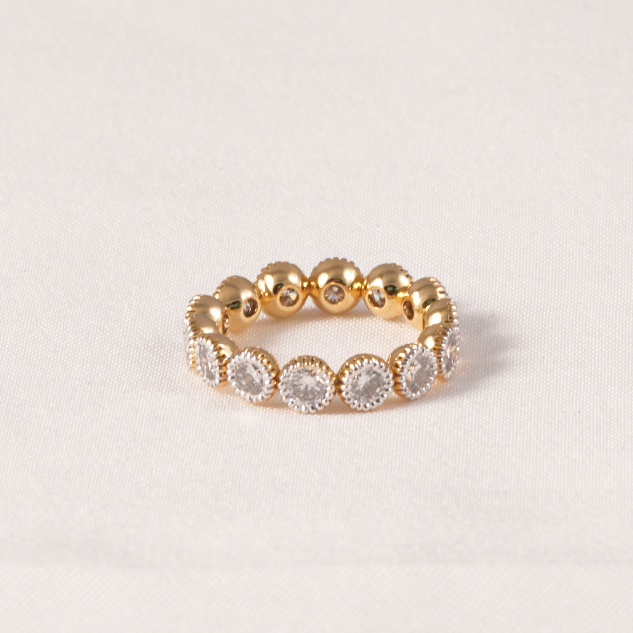 A gold beaded eternity ring featuring alternating smooth gold beads and oval clusters of clear round gemstones in a halo setting, displayed on a light beige textured fabric background.