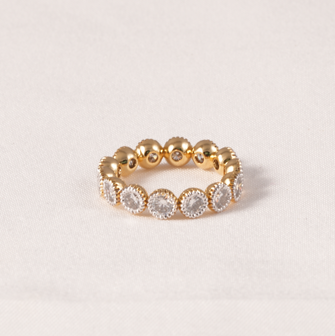 A gold beaded eternity ring featuring alternating smooth gold beads and oval clusters of clear round gemstones in a halo setting, displayed on a light beige textured fabric background.