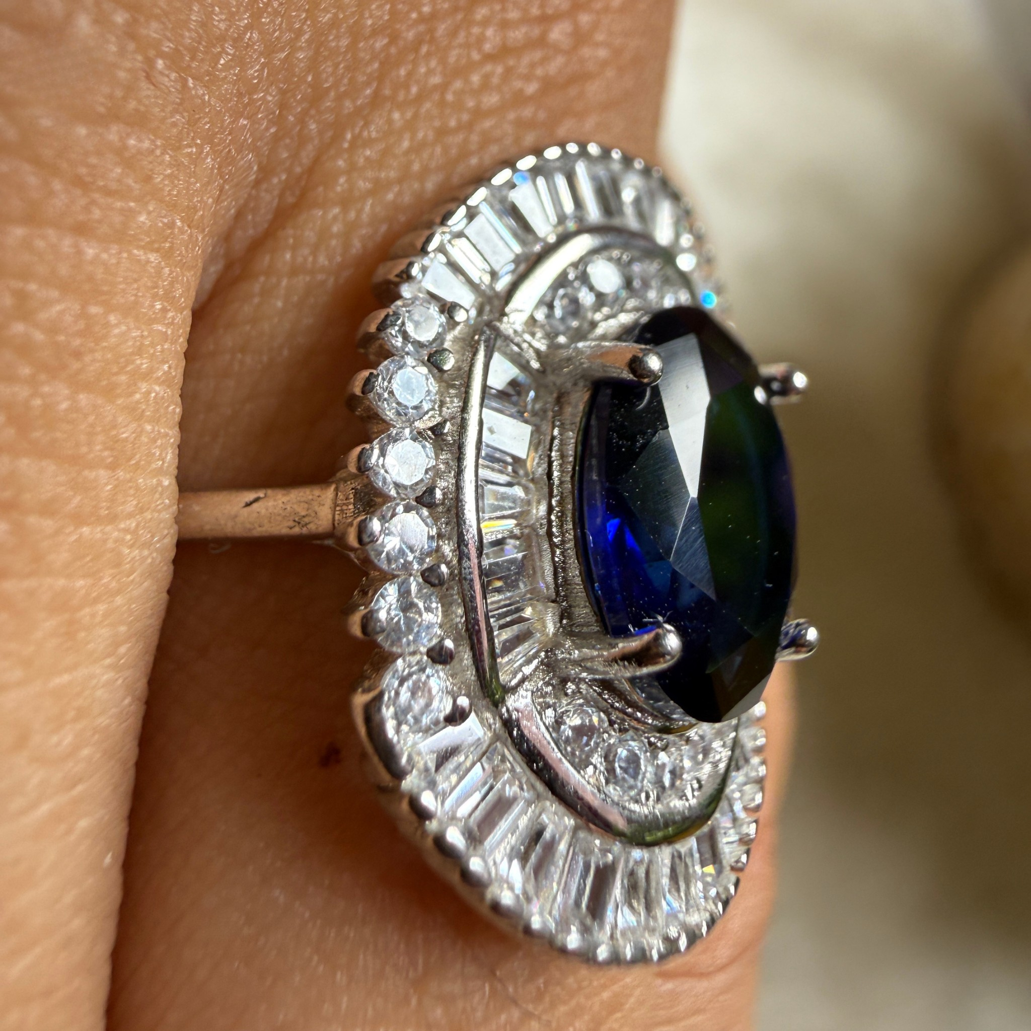 A close-up of a silver-toned halo ring with a dark blue oval center stone and clear baguette and round stones, shown worn on a finger.