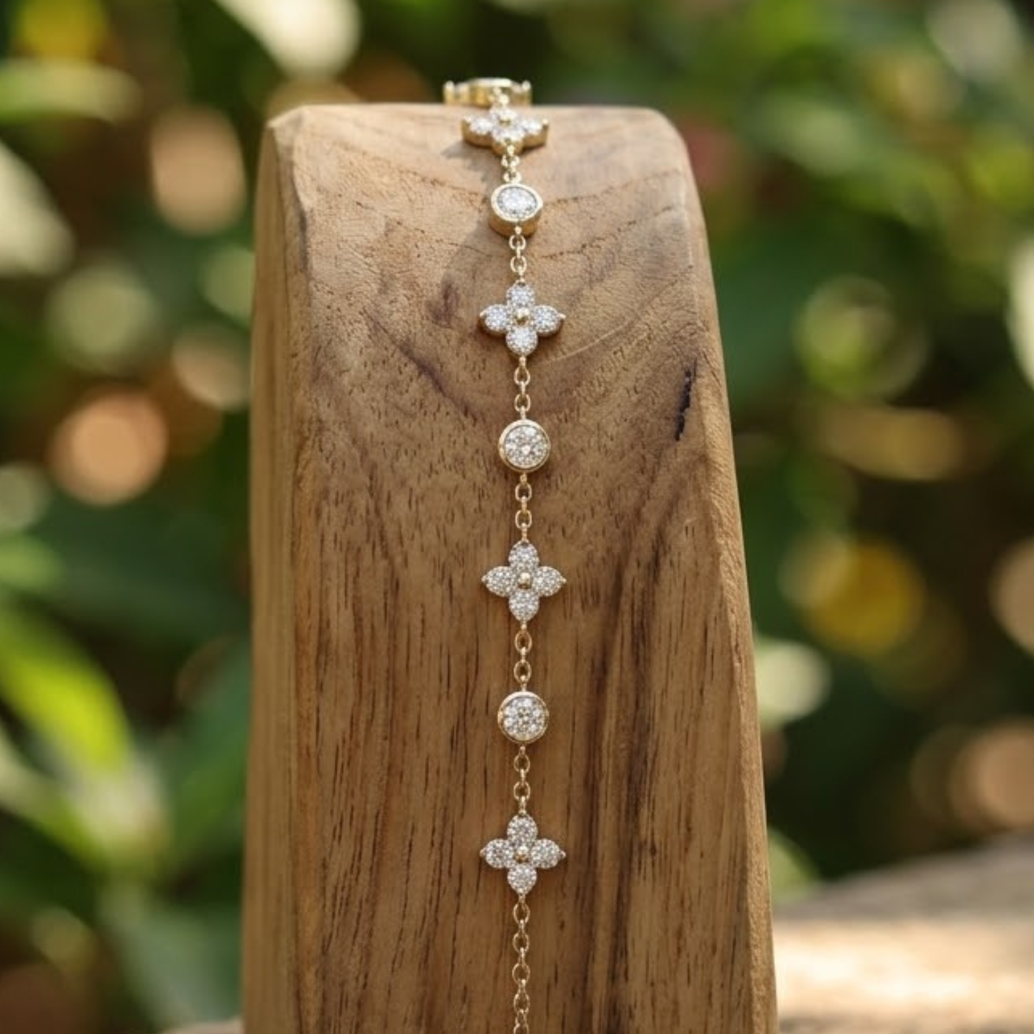 A delicate gold chain bracelet with alternating floral clover and round bezel-set clear stones, displayed on a rustic wooden block with a blurred green garden background.