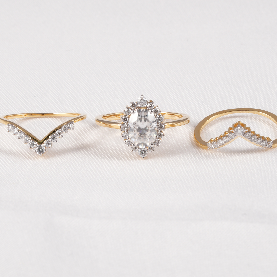Three gold rings displayed on a white fabric background: a V-shaped ring with small clear stones on the left, a halo-style ring with a large clear oval center stone in the middle, and a curved chevron ring with small clear stones on the right.