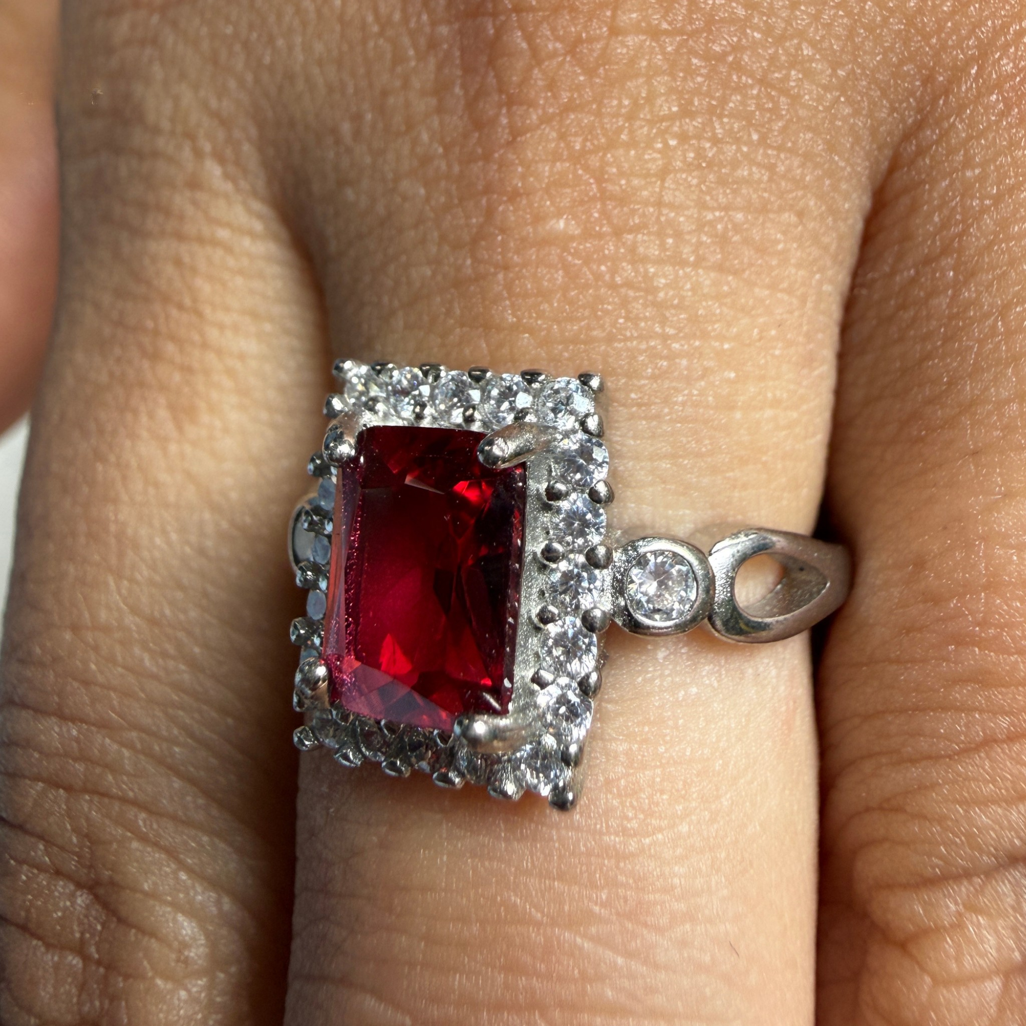A silver ring with a rectangular red gemstone surrounded by small clear stones, worn on a finger.