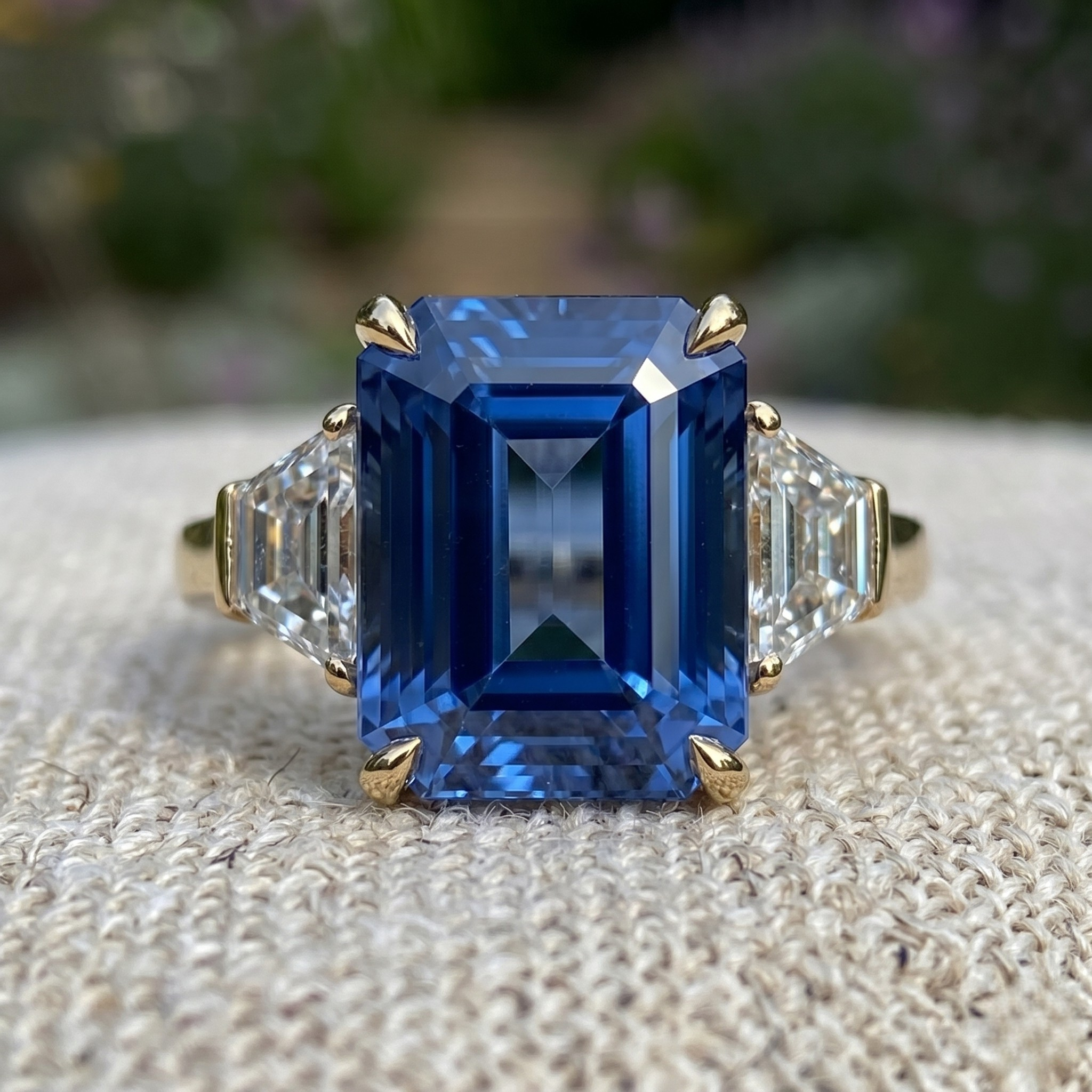 An emerald-cut blue sapphire ring set in yellow gold with baguette side stones, resting on a textured beige fabric with a blurred green garden background.