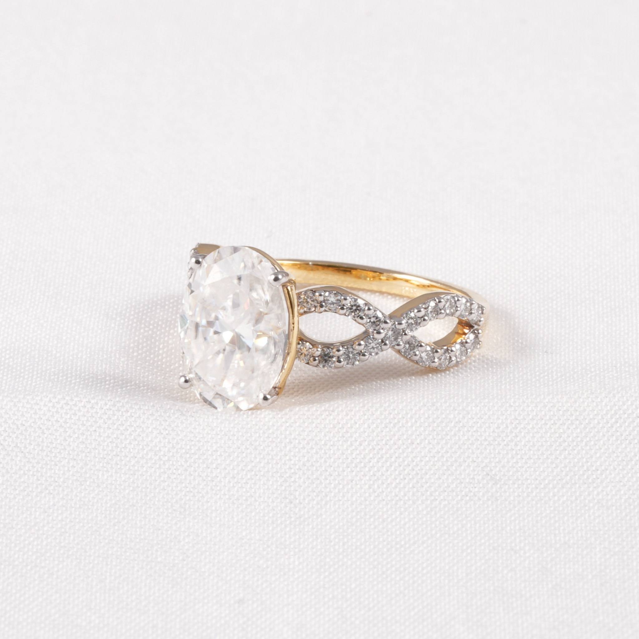A yellow gold ring featuring a large oval-cut clear gemstone in a prong setting, with an infinity-shaped band paved with small clear stones, shown on a textured white background.