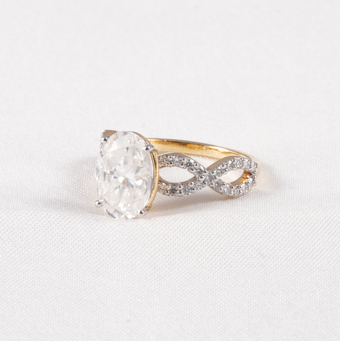 A yellow gold ring featuring a large oval-cut clear gemstone in a prong setting, with an infinity-shaped band paved with small clear stones, shown on a textured white background.