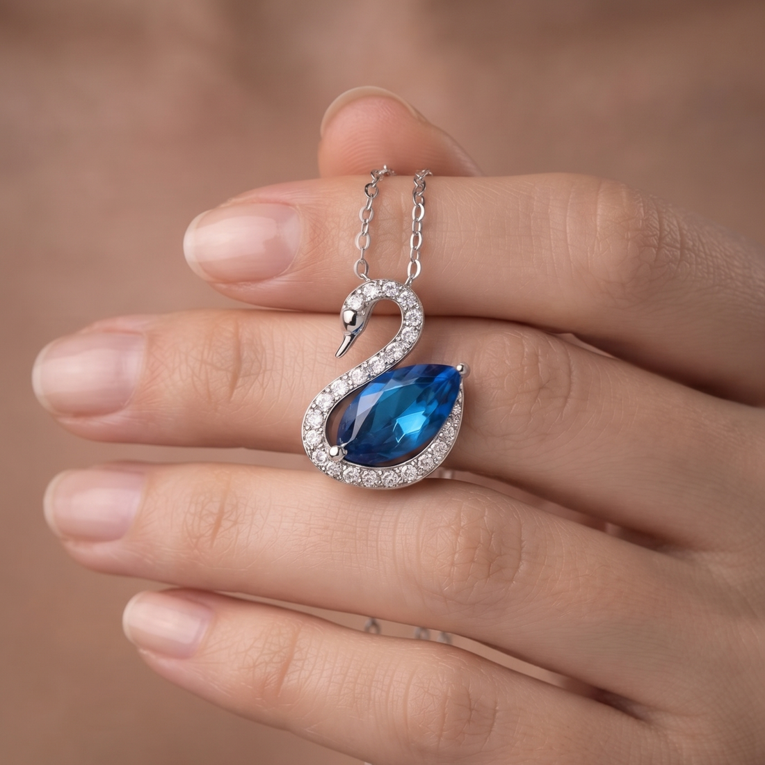 A silver-toned swan pendant necklace with a large blue teardrop gemstone and small clear stones, shown resting on a person's fingers against a soft warm brown background.