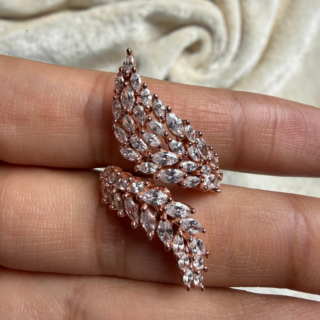 Rose gold leaf-shaped earrings with marquise-cut clear stones, shown worn on a person's fingers against a soft cream fabric background.