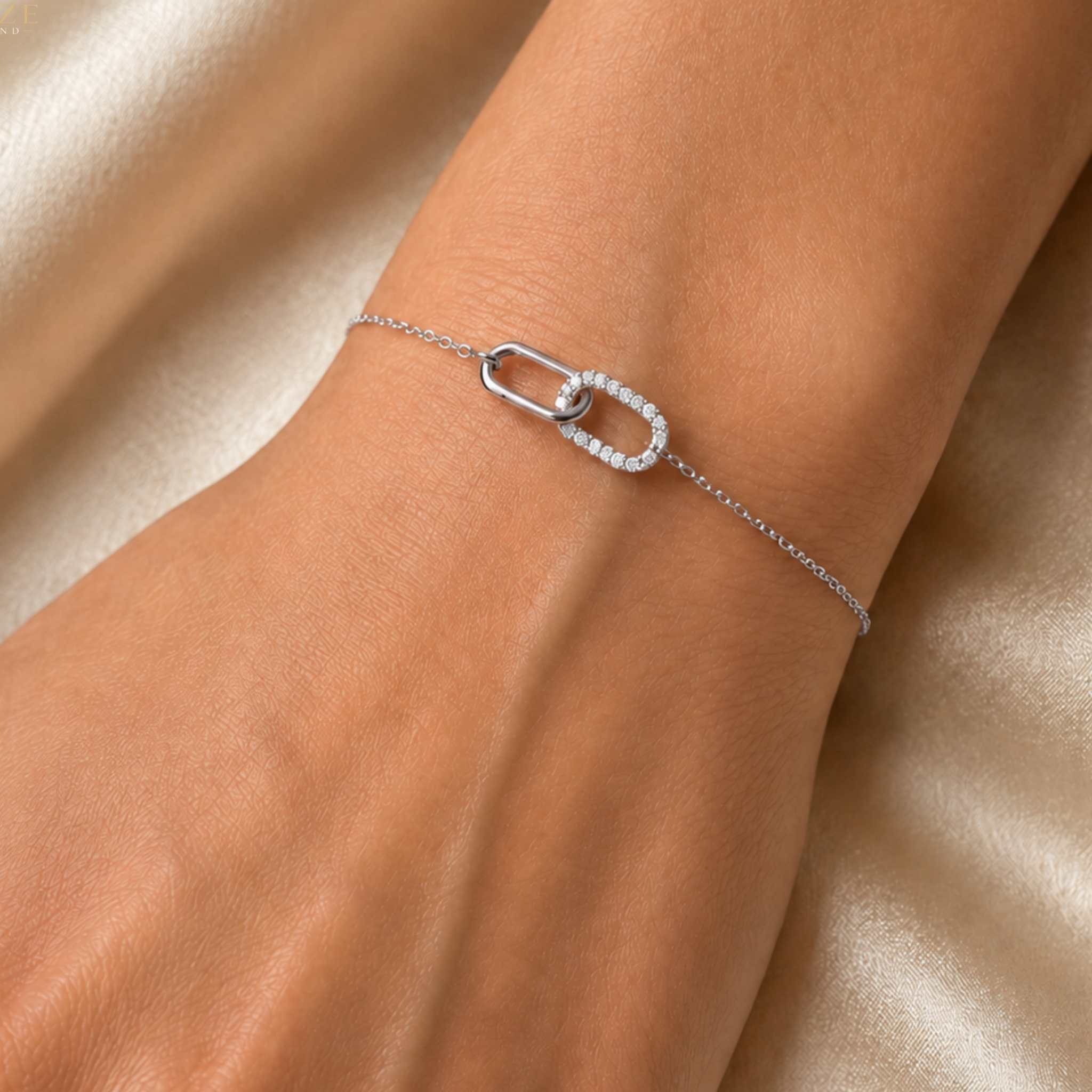 A delicate silver chain bracelet with a double interlocking oval link design, one of which is paved with small clear stones, shown worn on a wrist against a soft beige satin background.