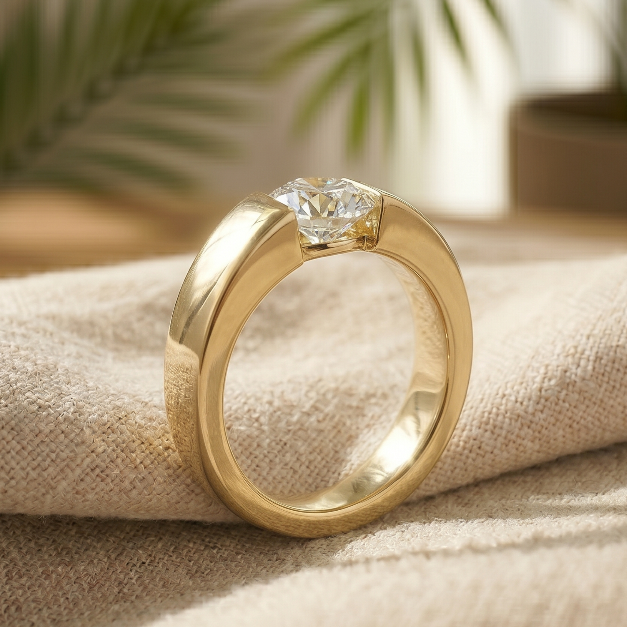 A polished yellow gold ring with a clear round-cut gemstone, resting on a textured beige linen fabric with a softly blurred background of green leaves.