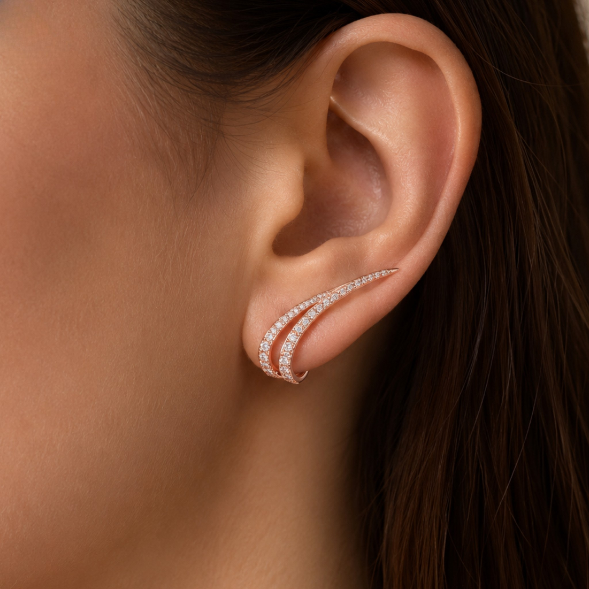 A close-up of a model's ear wearing a delicate rose gold ear cuff adorned with small sparkling clear stones.
