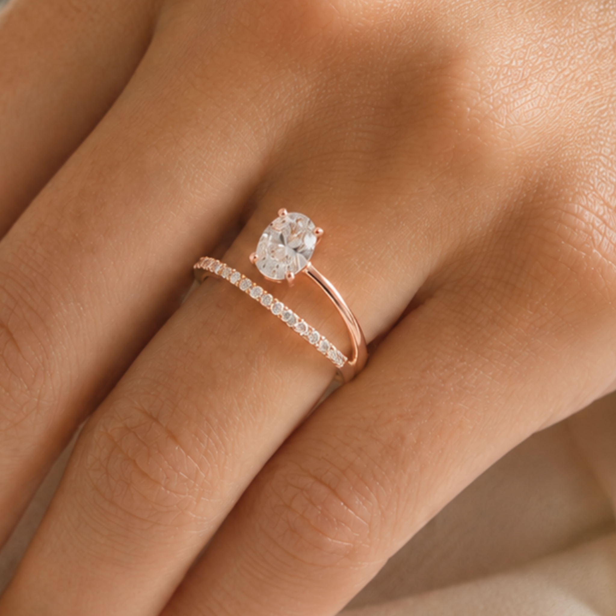 A close-up of a hand wearing a rose gold ring with a single oval-cut clear gemstone and a second thin pavé band ring.