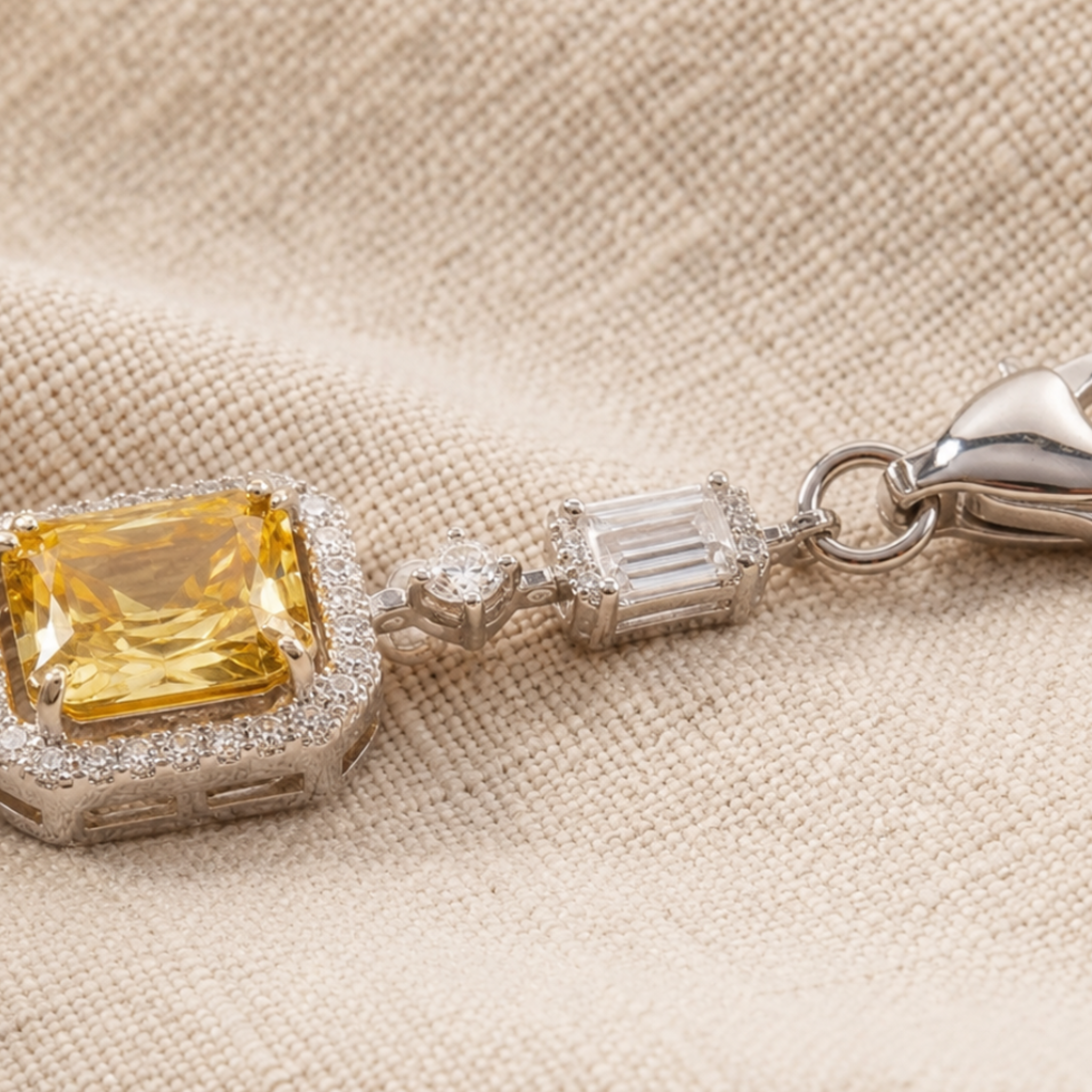 A close-up of a silver-toned jewelry piece featuring a large yellow cushion-cut gemstone surrounded by small clear stones, with a silver clasp, resting on a beige textured fabric background.
