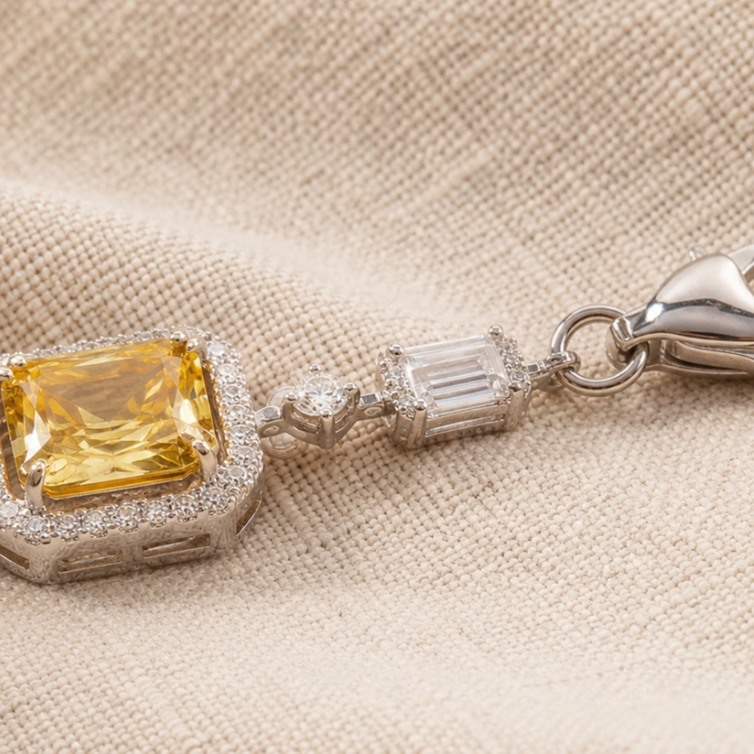 A close-up of a silver-toned jewelry piece featuring a large yellow cushion-cut gemstone surrounded by small clear stones, with a silver clasp, resting on a beige textured fabric background.