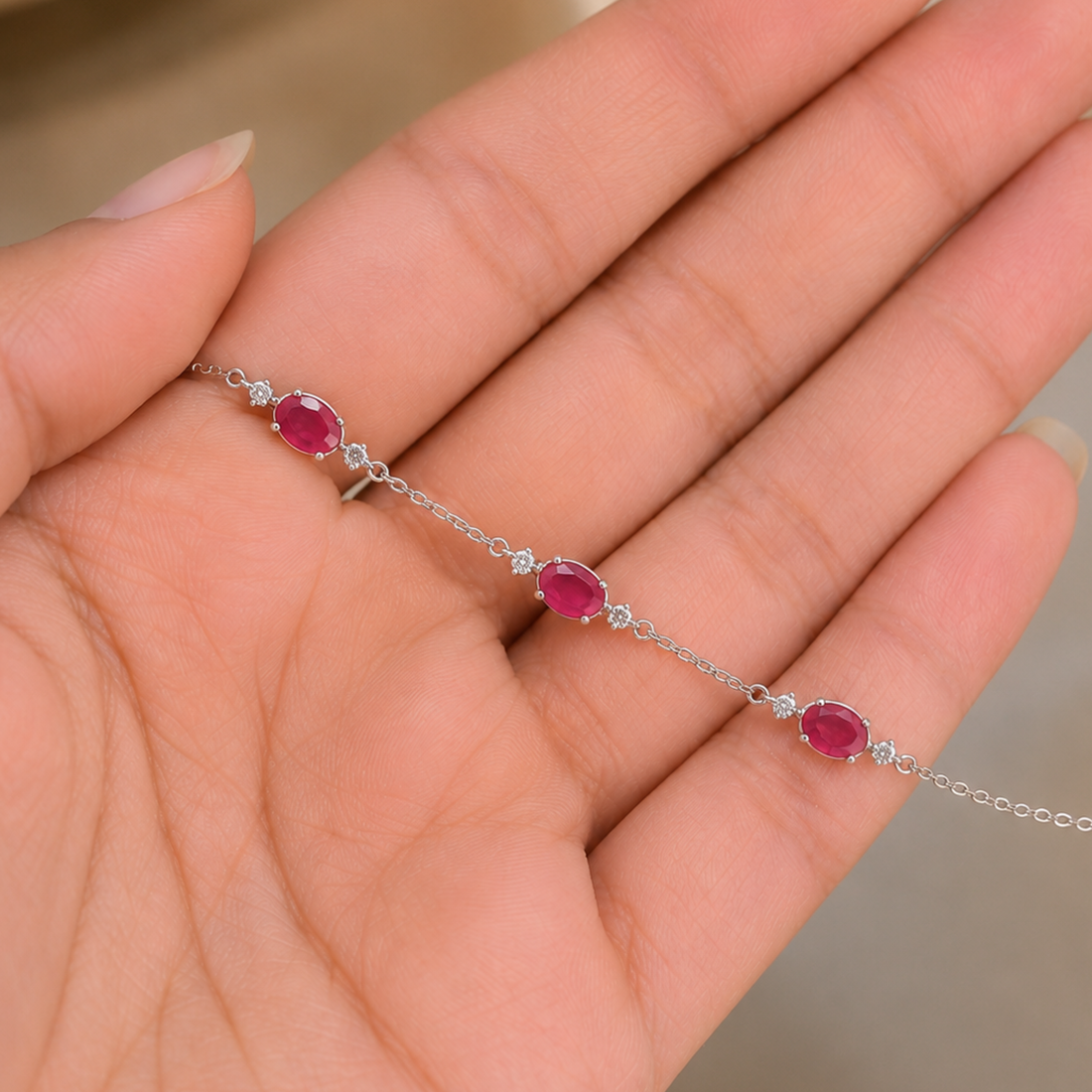 A delicate silver chain bracelet with three oval pinkish-red gemstones and small clear stones, shown resting on an open hand.
