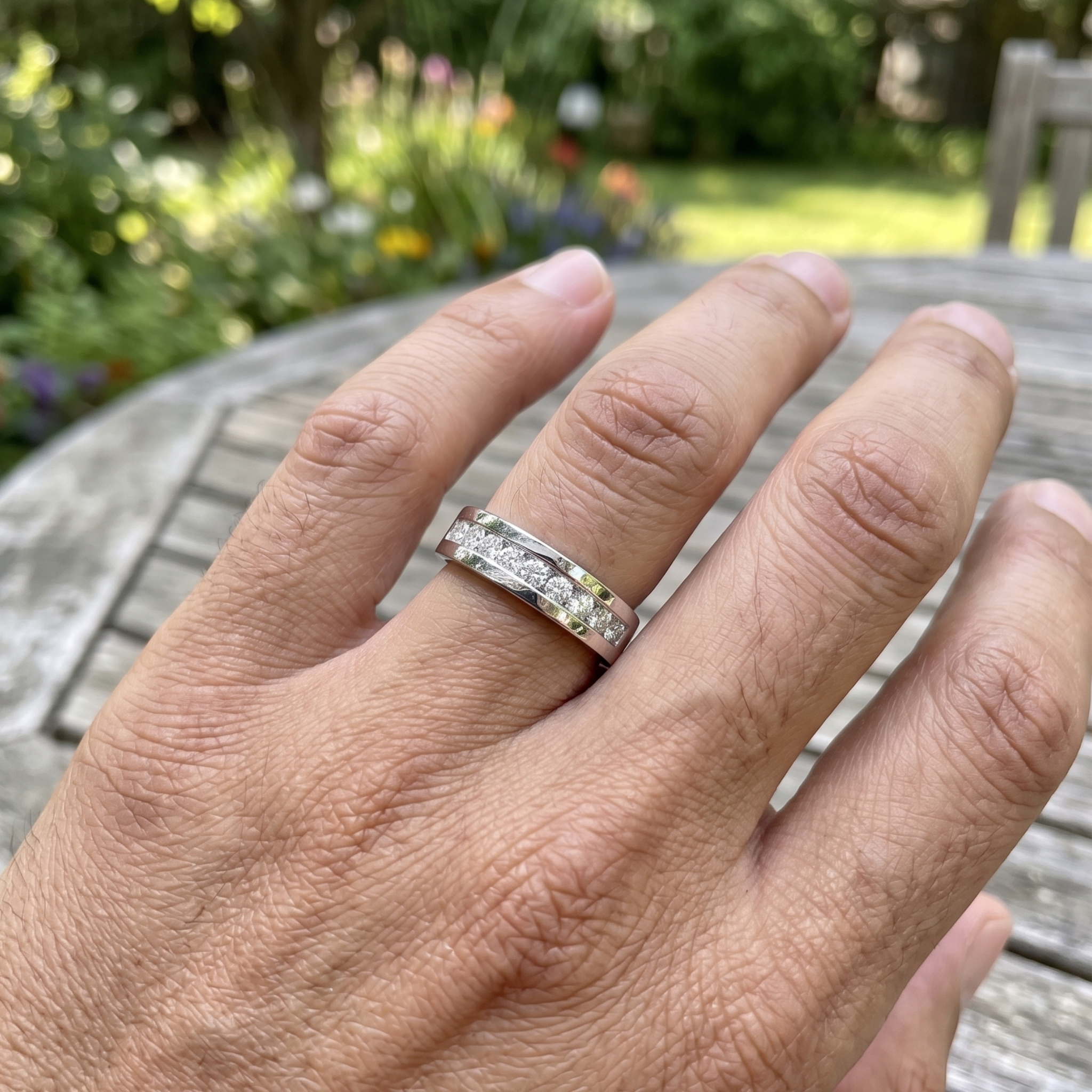 A silver band ring with small clear stones set along the top half, worn on a person's ring finger in an outdoor garden setting.