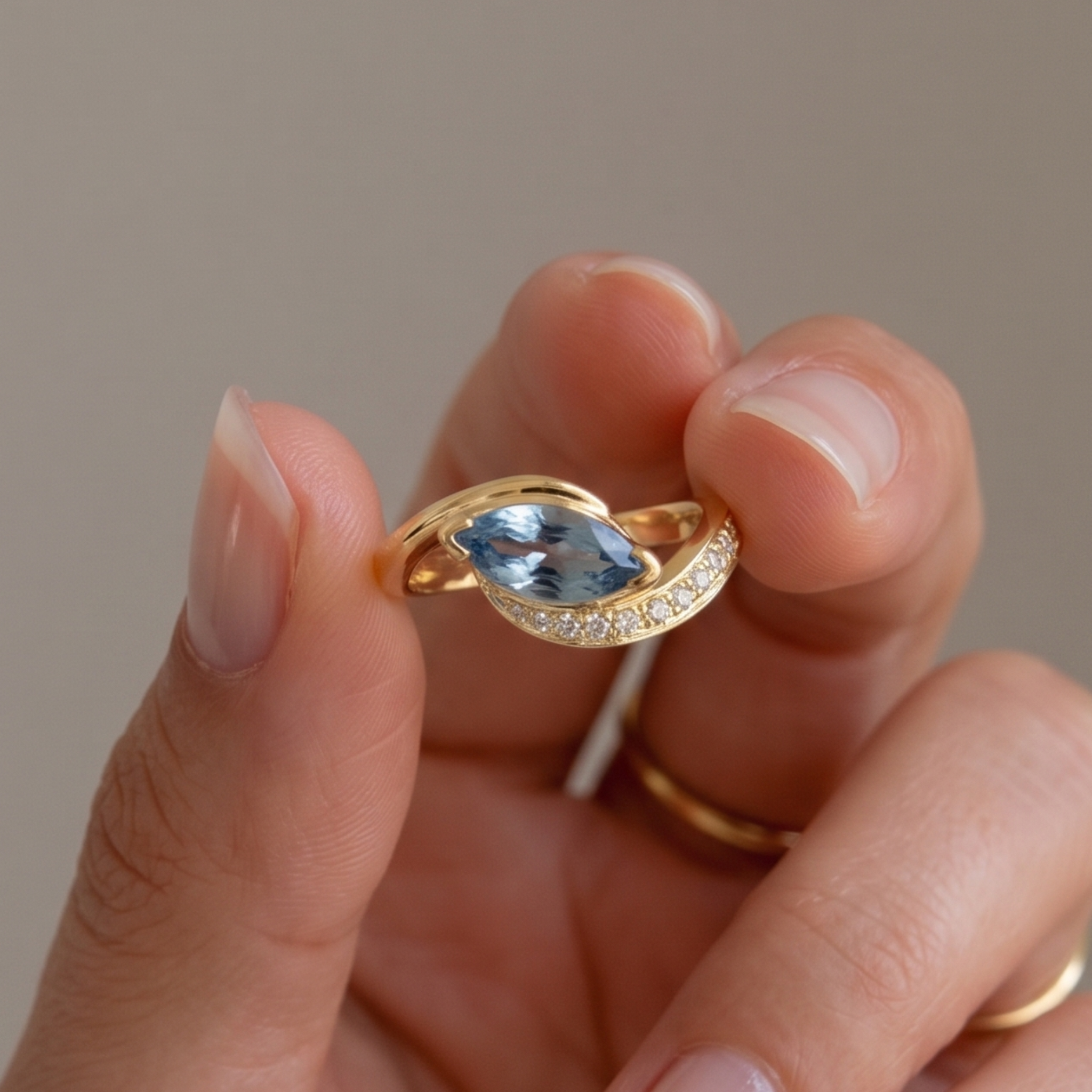 A close-up of a hand holding a gold ring with a marquise-cut light blue gemstone and a curved row of small clear stones on the band, against a soft neutral background.