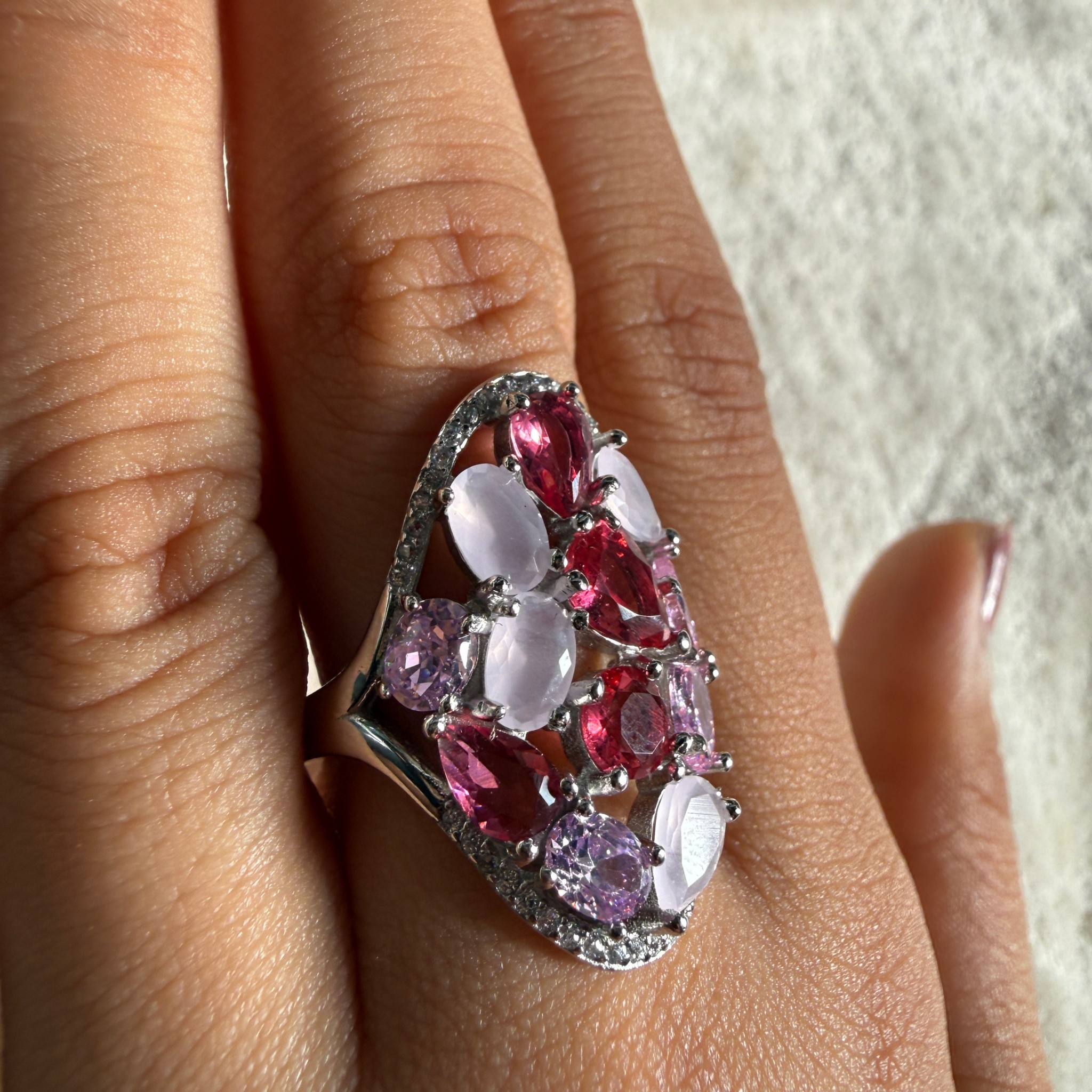 A close-up of a hand wearing a large statement ring. The ring features multiple oval-cut pink and purple gemstones with clear stones, set in a silver-toned metal.