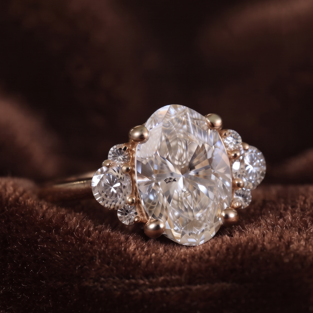 An oval-cut diamond ring with a rose gold setting, accented by smaller round diamonds, resting on a textured brown velvet surface.