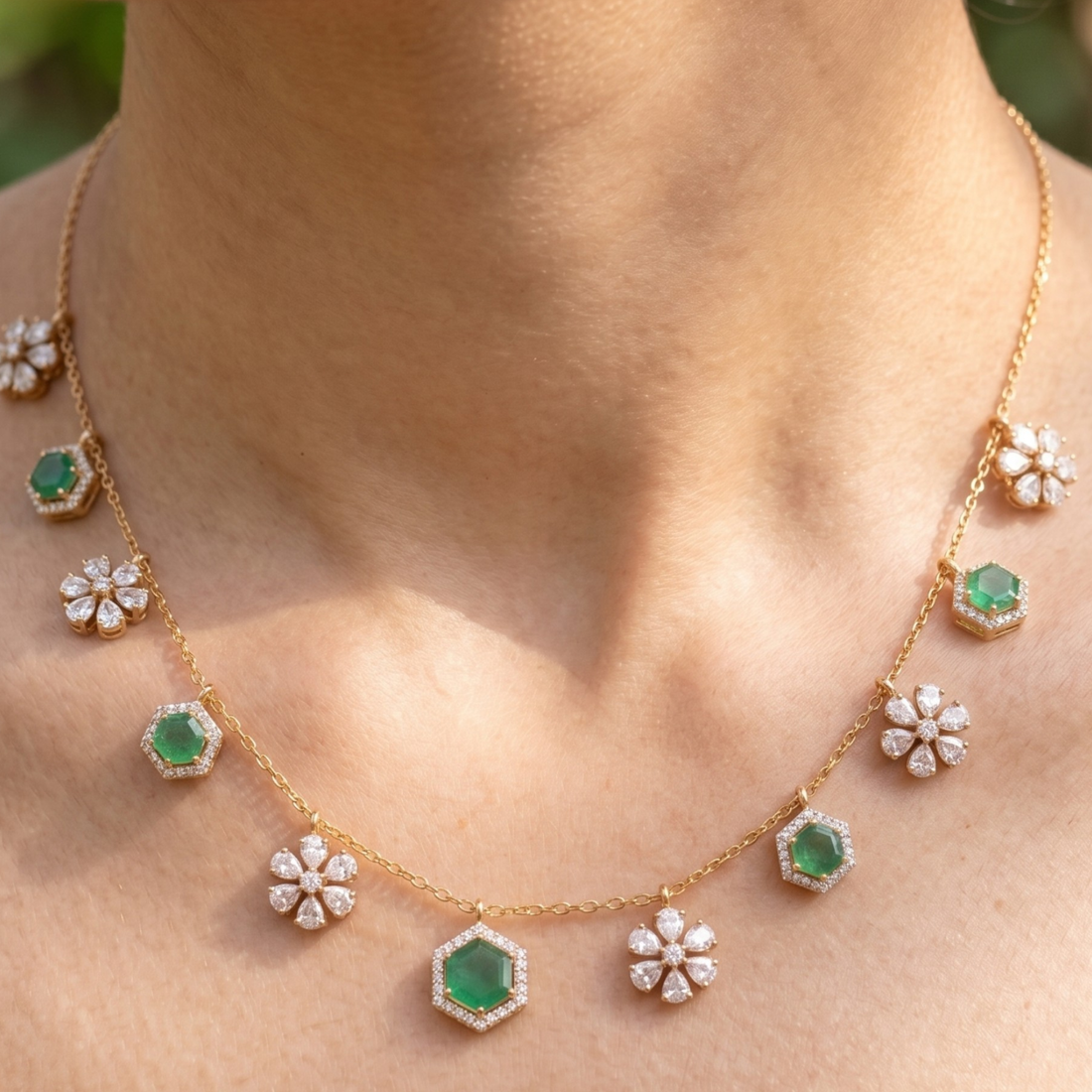 A gold necklace with green emerald-cut stones and white flower-shaped pavé accents, shown worn on a person's neck.