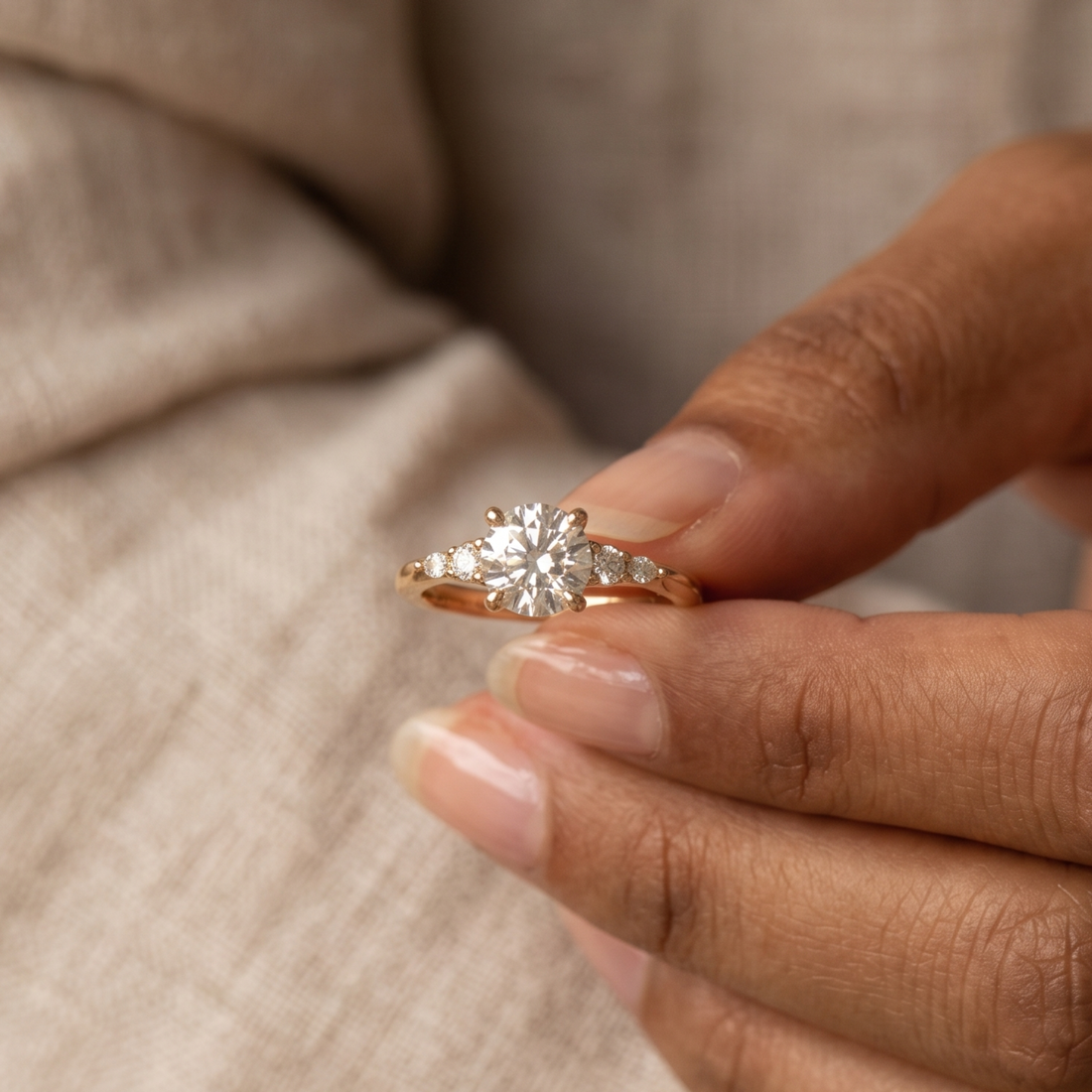 A close-up of a hand holding a rose gold ring with a central round diamond and small side diamonds, set against a soft beige textured fabric background.