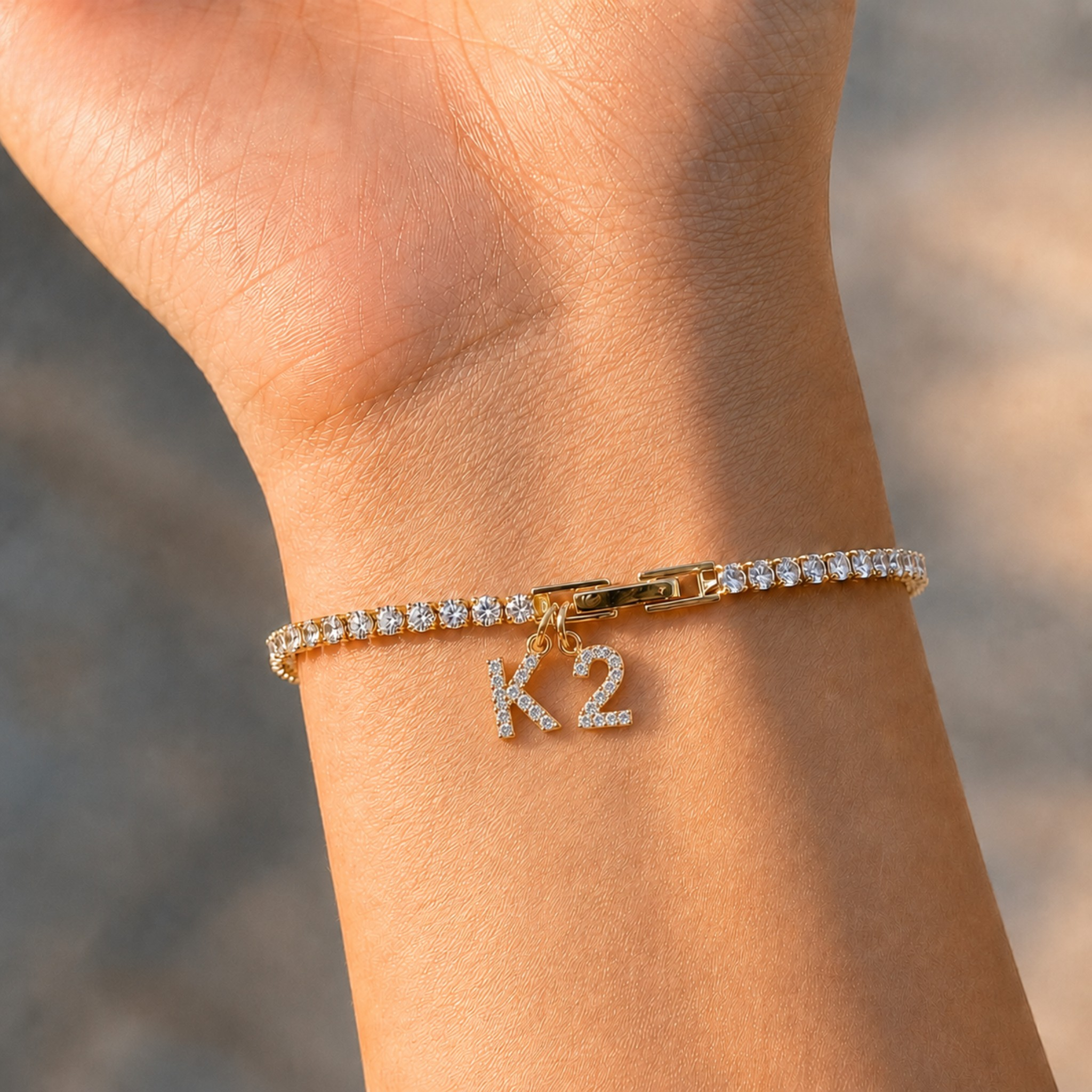A gold bracelet with a row of clear pavé stones and a dangling 'K2' initial charm, shown worn on a person's wrist.